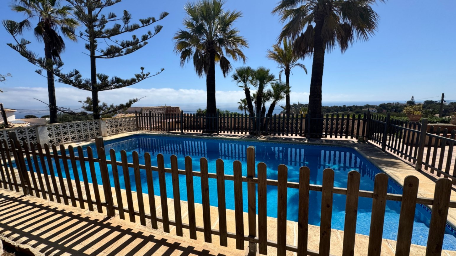 Piscine extérieure avec mosaïque bleue, entourée d'une clôture en bois. L'espace comprend des palmiers et offre une vue dégagée sur le littoral méditerranéen.