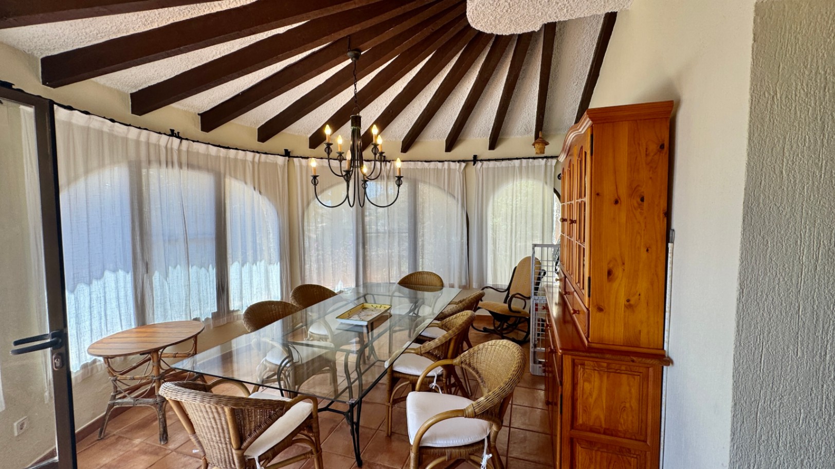 Salle à manger avec plafond à poutres rayonnantes, table en verre, chaises en osier et buffet en bois massif sur un sol en terre cuite.