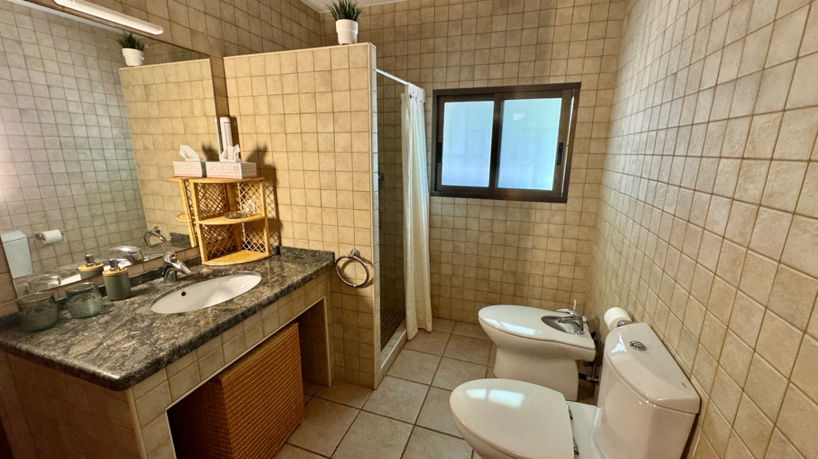 Salle de bain avec plan de travail en granit, vasque intégrée et rangements en osier. Murs carrelés en beige, douche à l'italienne avec rideau et espace bidet.