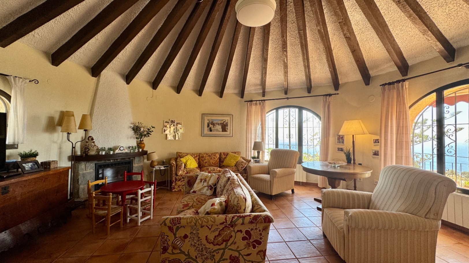 Salon circulaire avec plafond voûté à poutres de bois radiales, sol en terre cuite, cheminée en pierre et fenêtres en arc offrant une vue sur la mer.