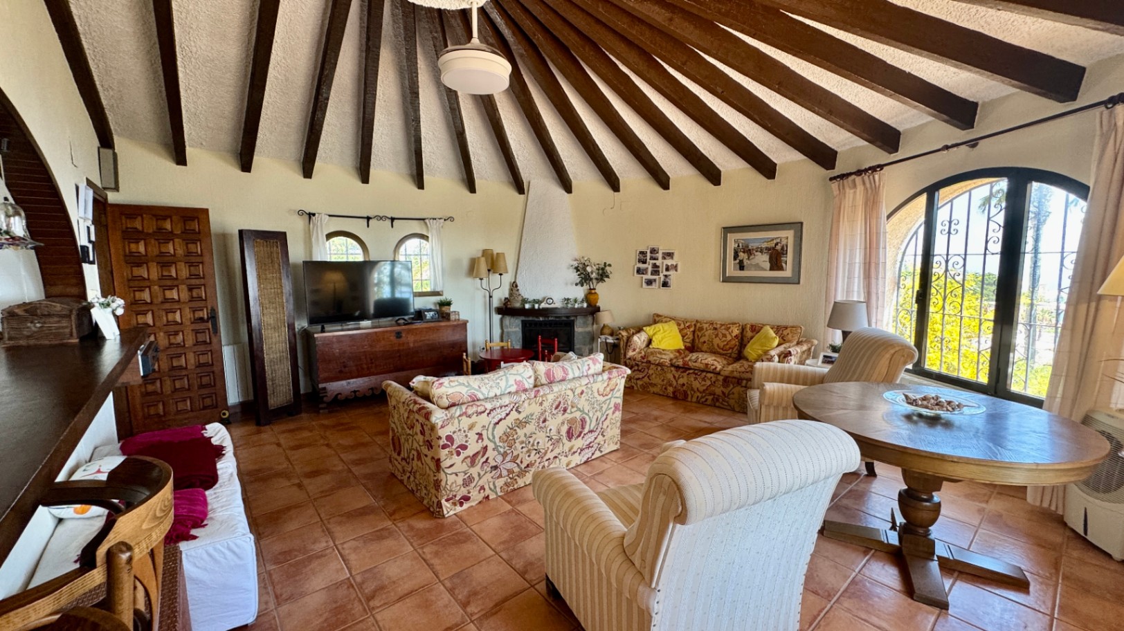 Salon spacieux avec plafond voûté à poutres apparentes radiales, sol en terre cuite, cheminée et fenêtres cintrées avec ferronnerie décorative.