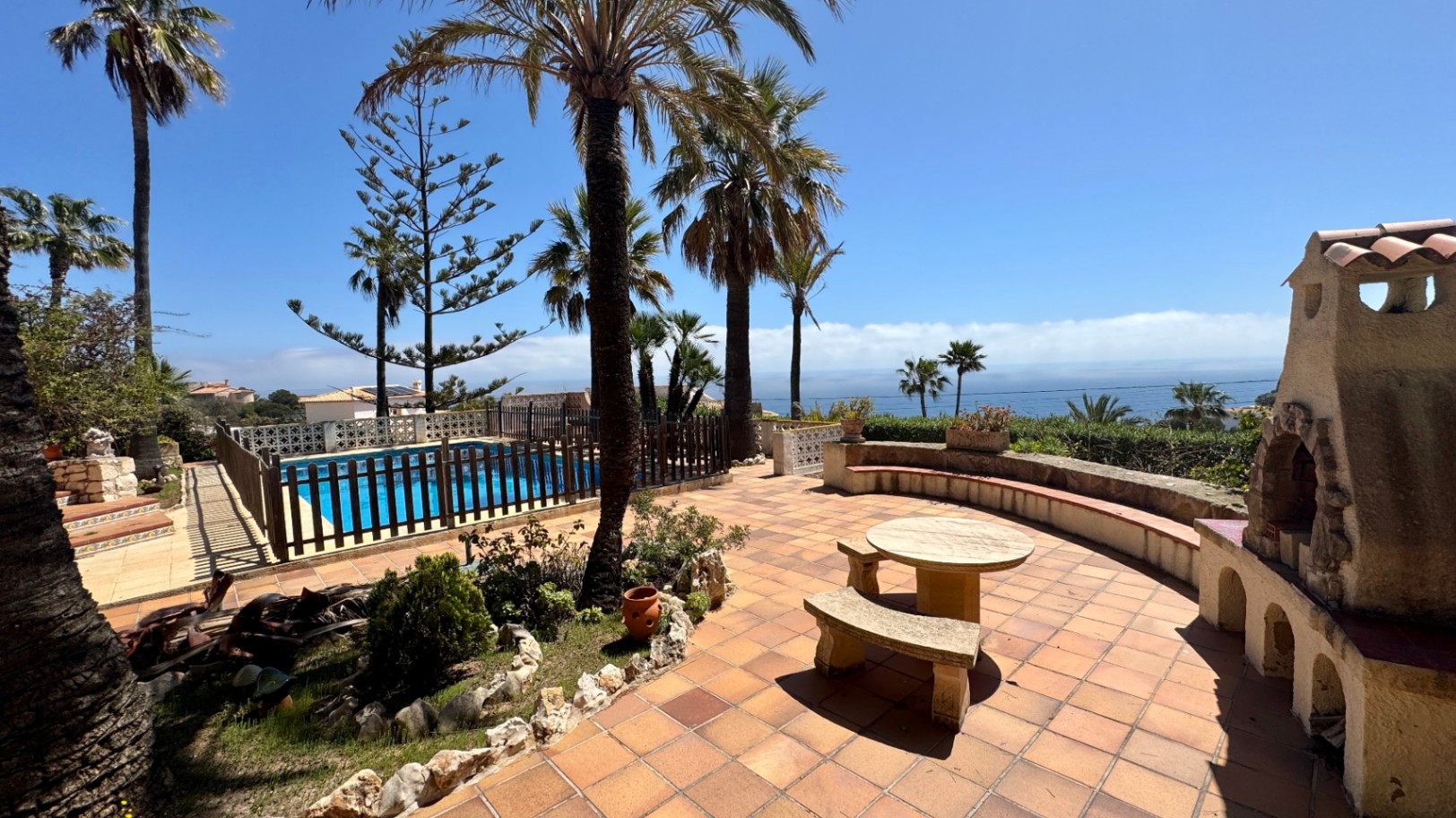 Terrasse en terre cuite avec table en pierre, cheminée extérieure rustique et piscine clôturée offrant une vue dégagée sur la mer et les palmiers.