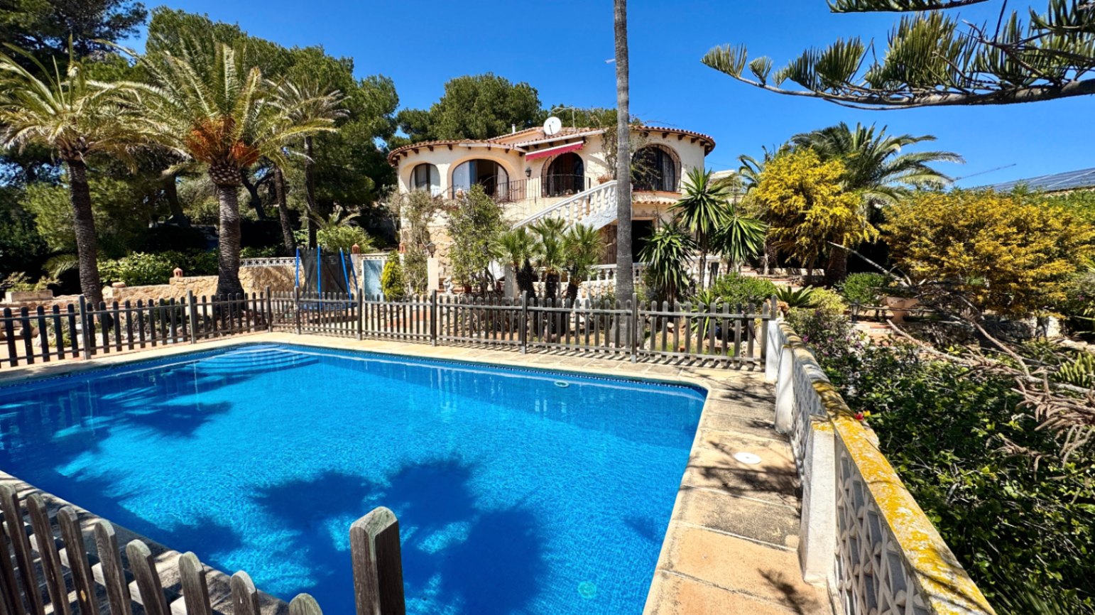 Vue extérieure d'une piscine rectangulaire clôturée devant une villa méditerranéenne aux balcons cintrés, entourée de palmiers et de pins.