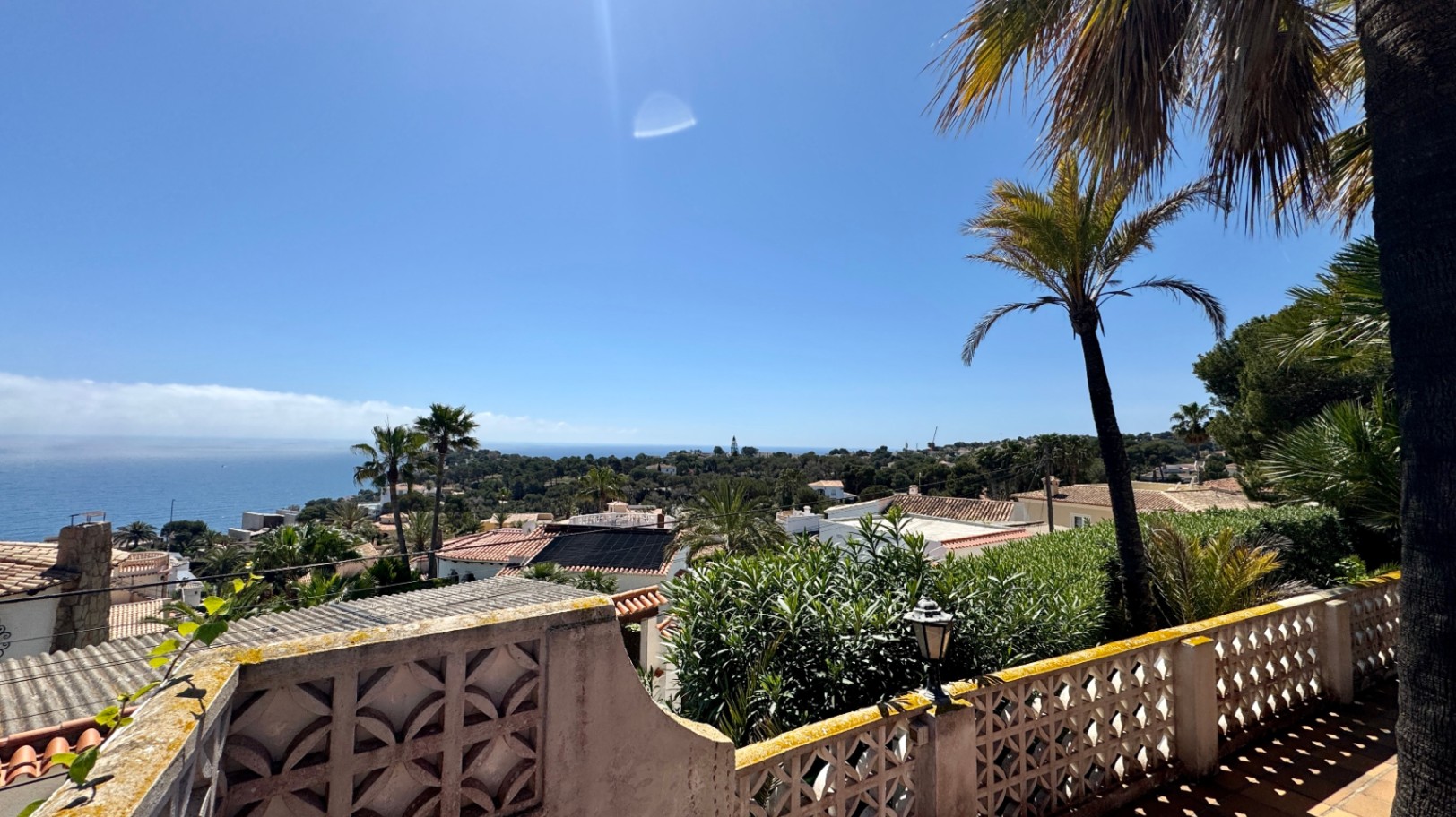 Vue surélevée depuis une terrasse avec balustrade sculptée, surplombant des toits en terre cuite, des palmiers et l'horizon marin sous un ciel dégagé.