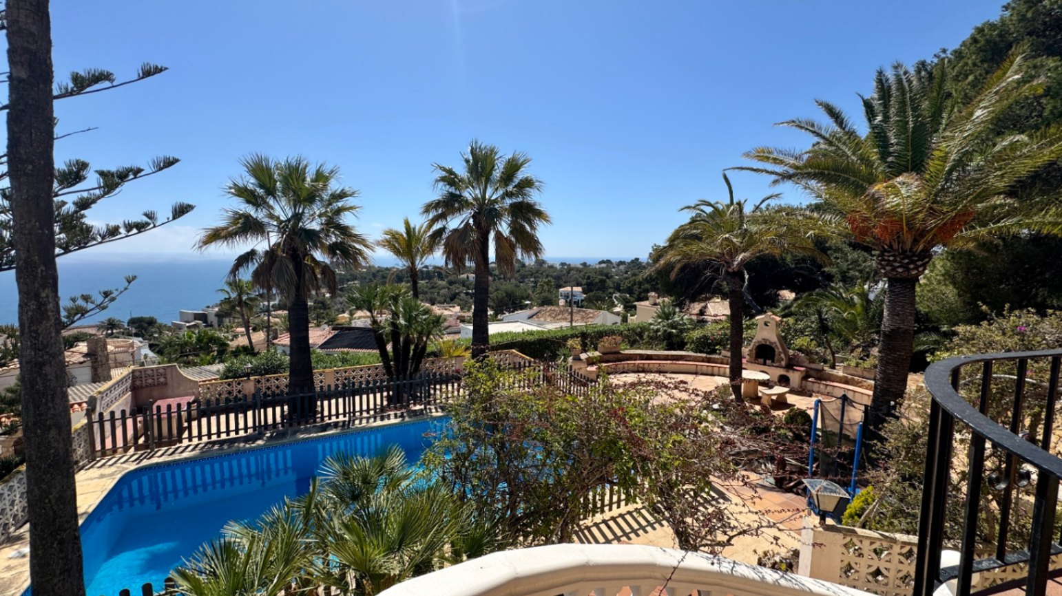 Vue surélevée d'une piscine privée et d'une terrasse en pierre avec cheminée extérieure, entourée de palmiers et offrant une vue sur la mer Méditerranée.