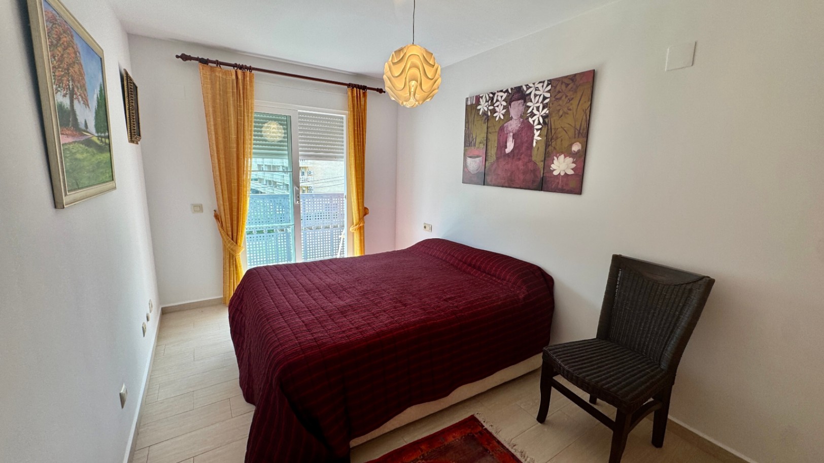 Chambre avec lit double, couvre-lit bordeaux et chaise en osier. Une porte coulissante vitrée avec rideaux jaunes apporte de la luminosité et donne sur le balcon.