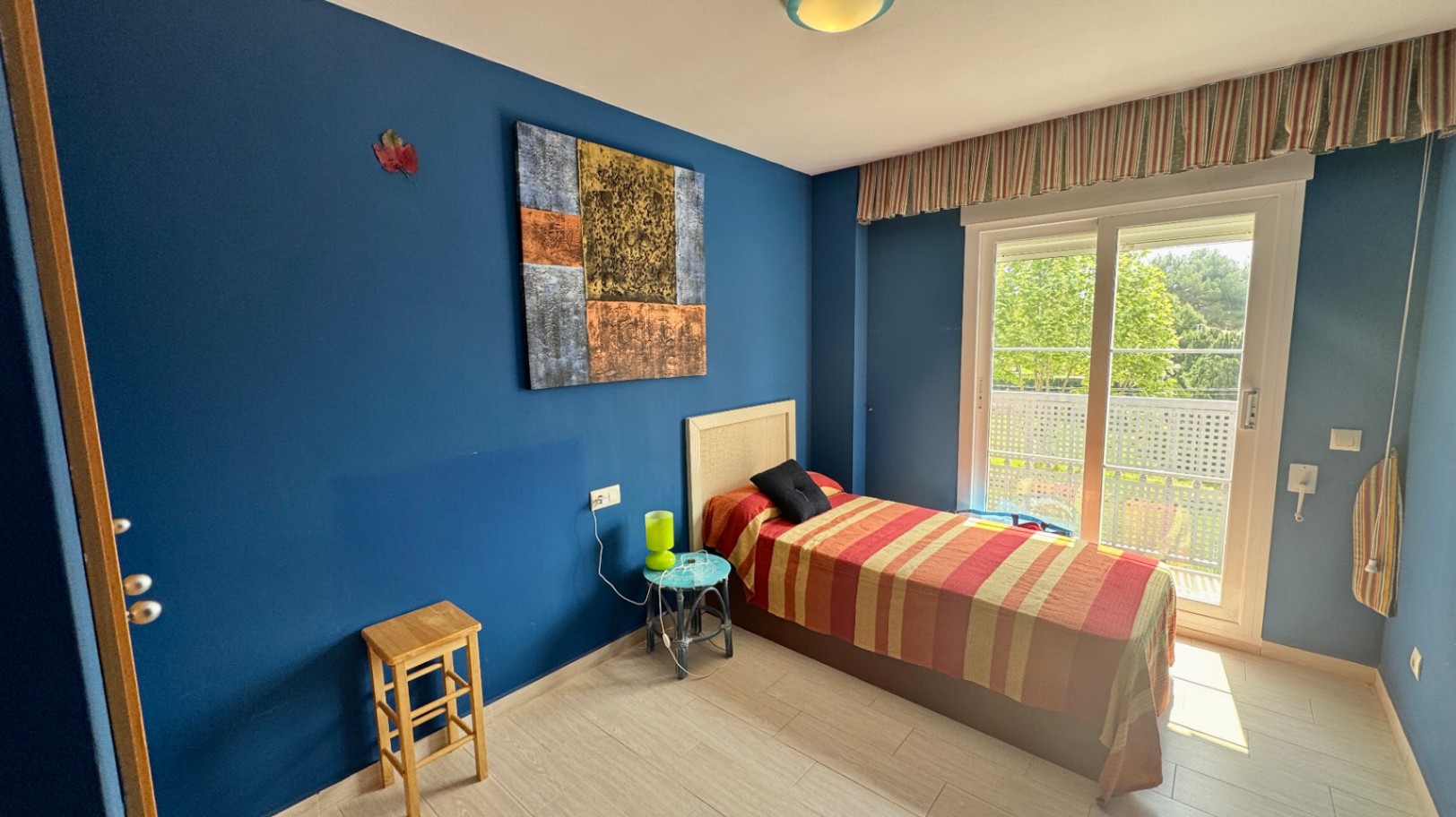 Chambre individuelle aux murs bleu profond, dotée d'une tête de lit en osier et d'une grande baie vitrée ouvrant sur un balcon avec vue dégagée.