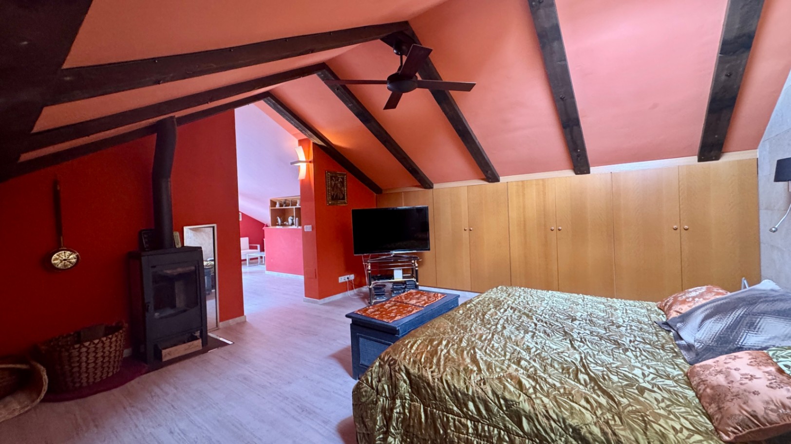 Chambre mansardée avec poutres apparentes, murs terracotta, poêle à bois en fonte et placards intégrés sur toute la longueur en bois clair.