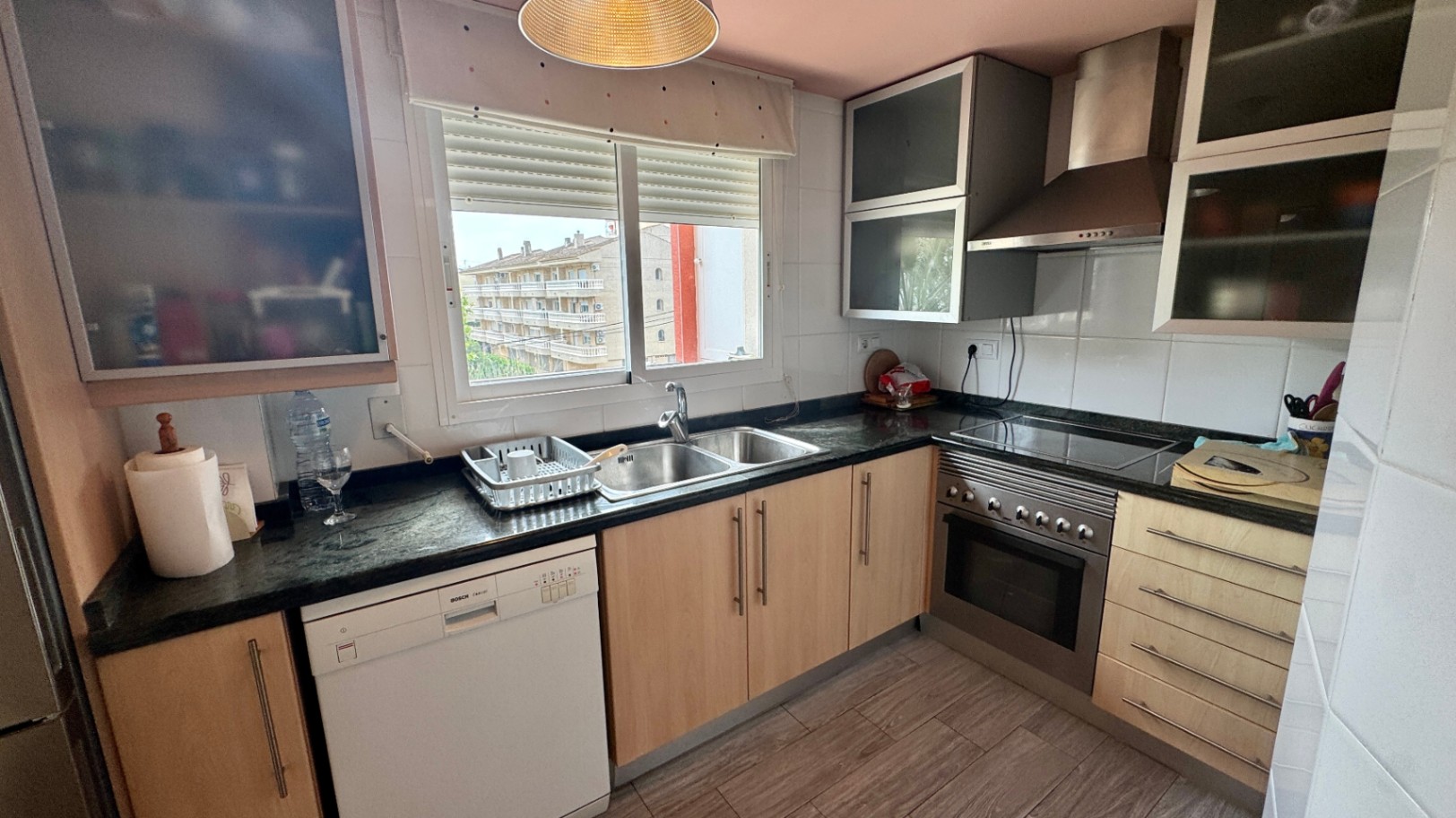 Cuisine fonctionnelle avec placards en bois clair, plan de travail en granit sombre, lave-vaisselle Bosch et évier double face à une fenêtre.