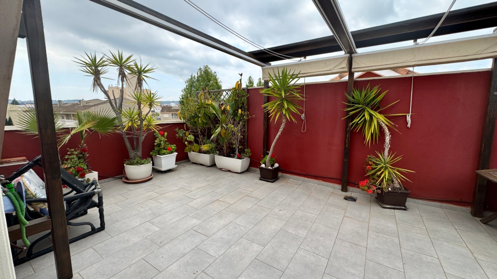 Grande terrasse de toit avec carrelage gris, pergola en bois munie de stores rétractables et diverses plantes en pots disposées le long d'un mur rouge bordeaux.