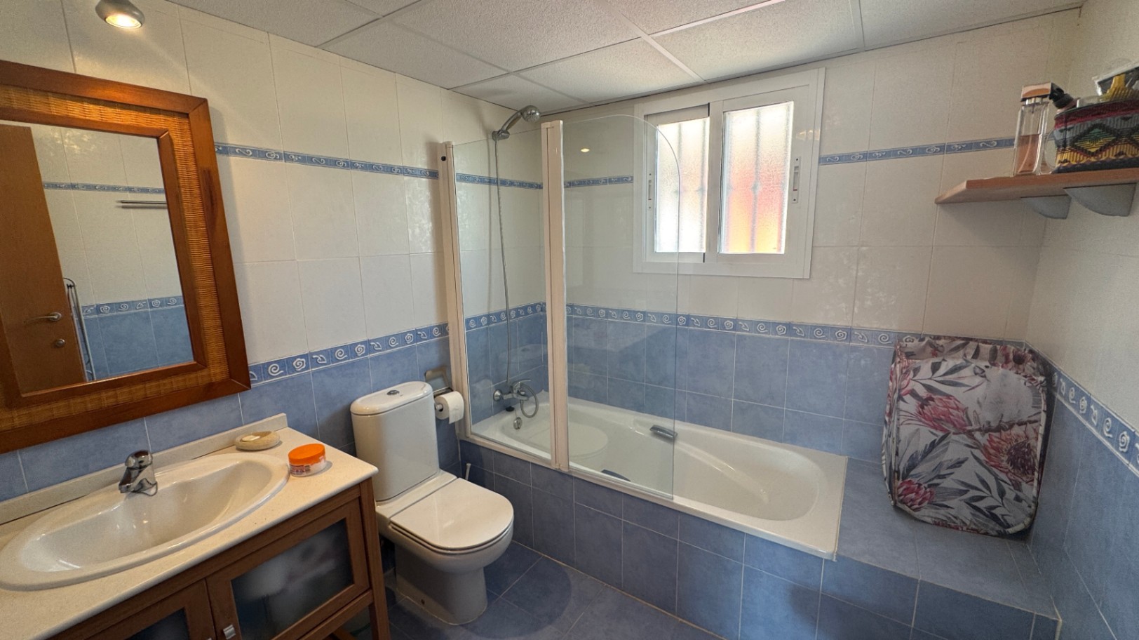 Salle de bain avec carrelage bleu et blanc, baignoire équipée d'un pare-douche en verre, miroir avec cadre en bois et meuble vasque assorti.