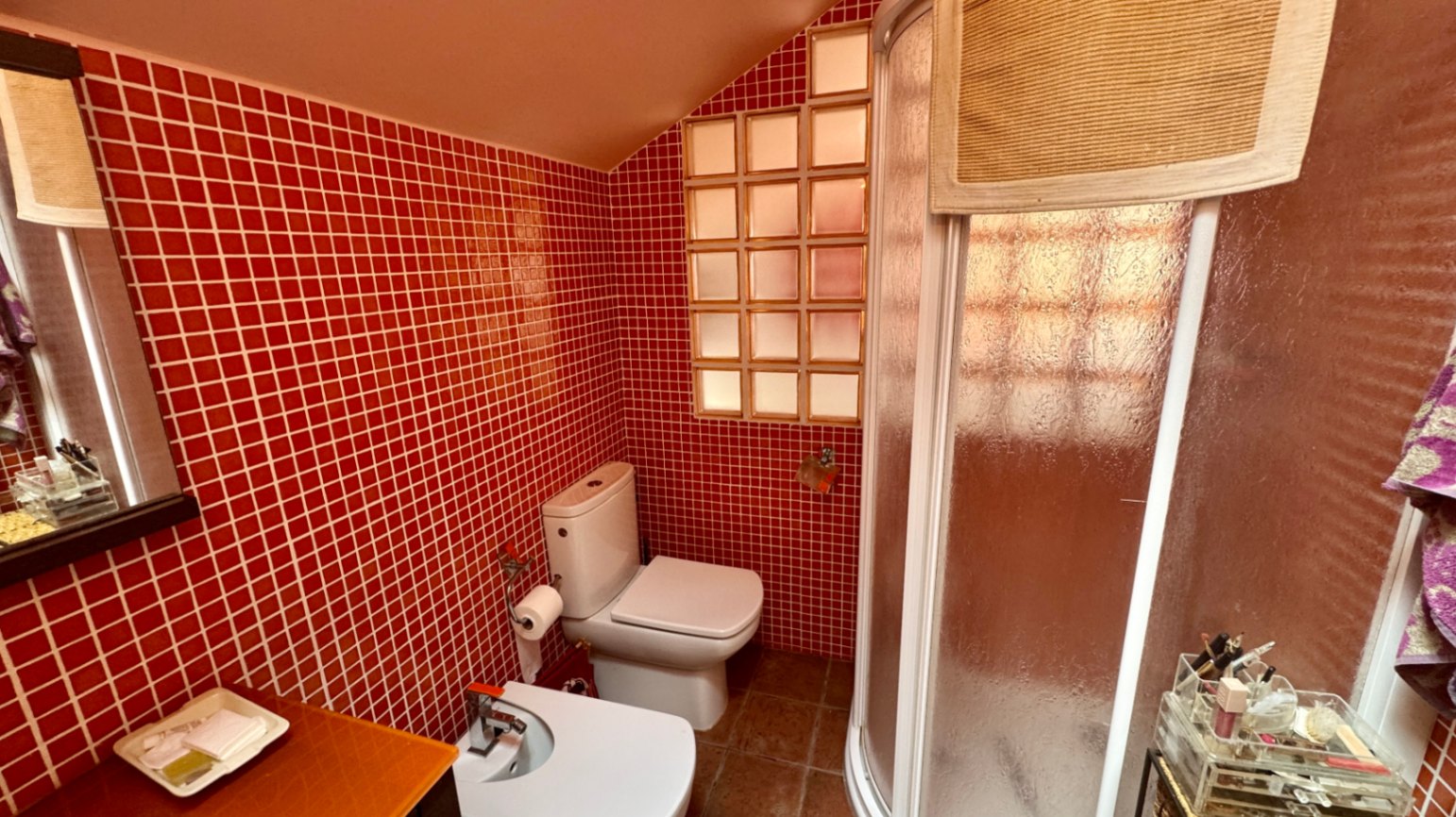 Salle de bain avec murs en mosaïque rouge, plafond mansardé et douche d'angle courbée. Elle comprend un WC, un bidet et une fenêtre en briques de verre.