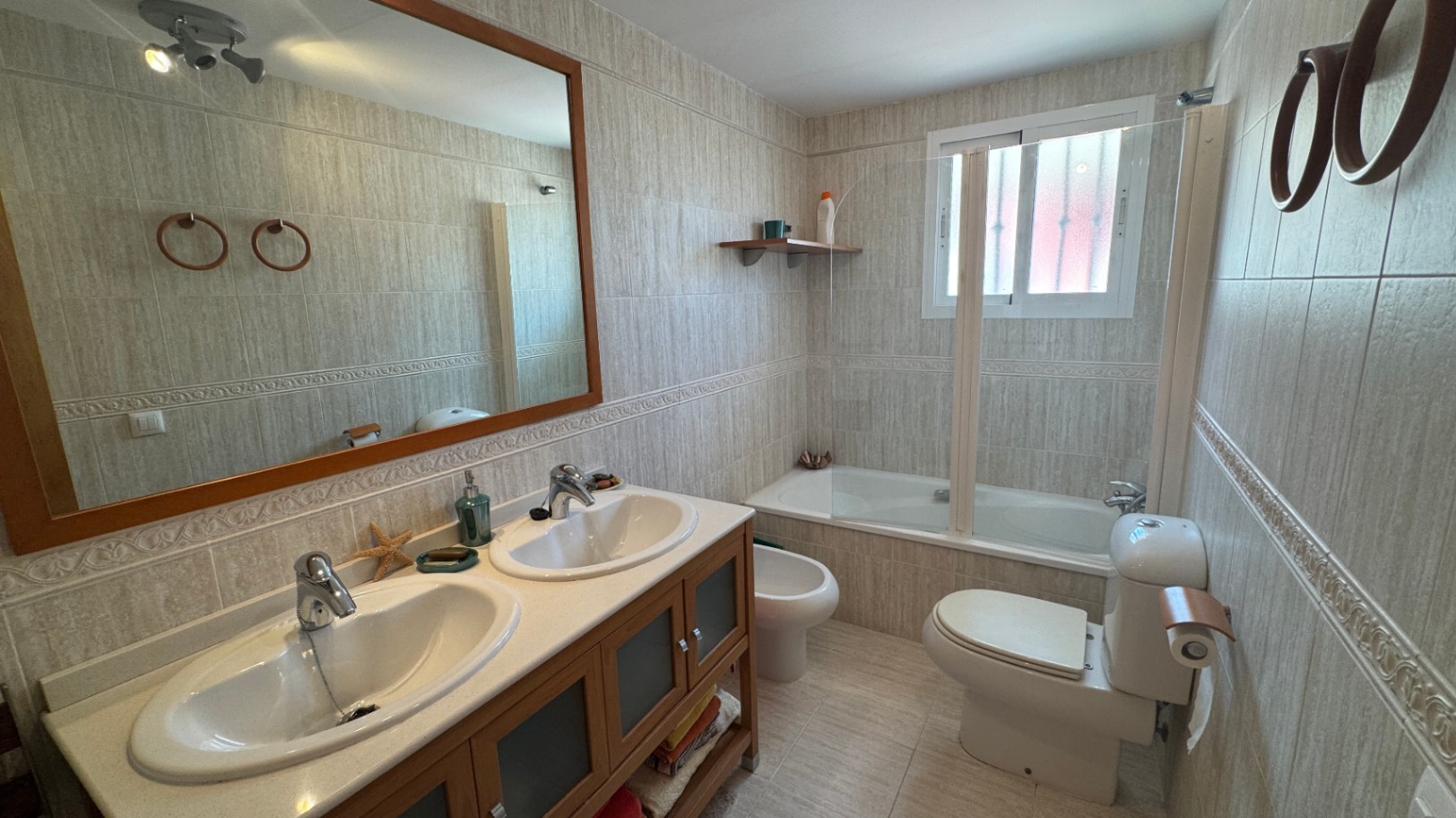 Salle de bain équipée d'un double lavabo avec meuble en bois, d'une baignoire avec paroi vitrée et de murs carrelés dans des tons naturels.