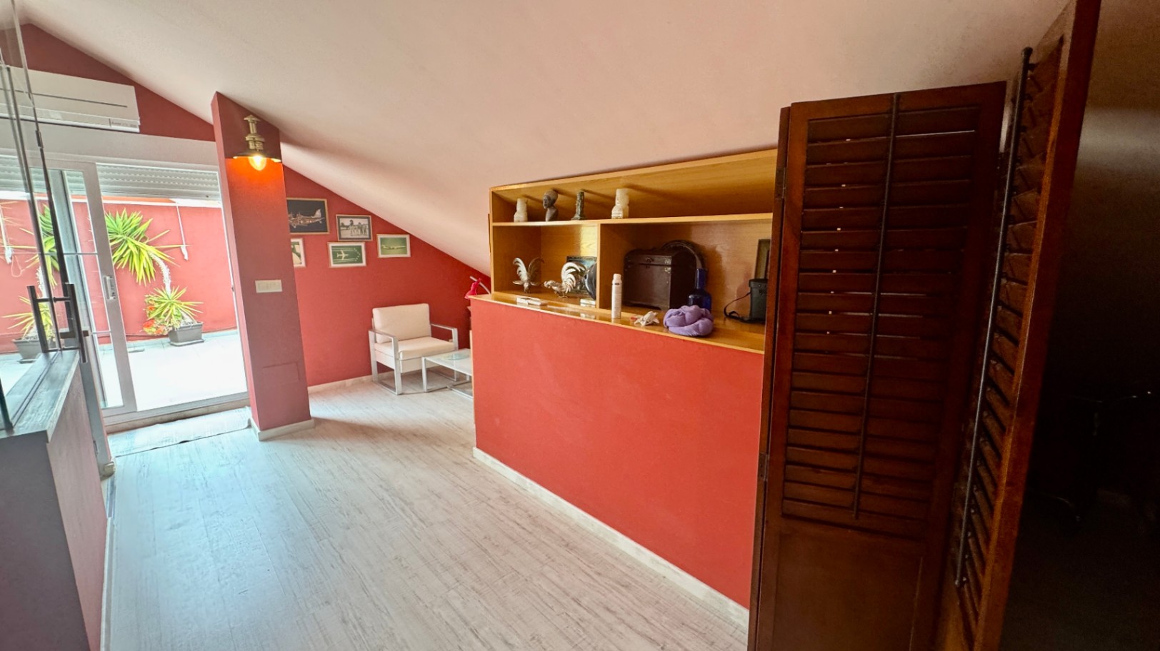 Salon mansardé aux murs terracotta avec étagères en bois intégrées et plafonds inclinés. Une baie vitrée s'ouvre sur une terrasse privée avec des plantes en pot.