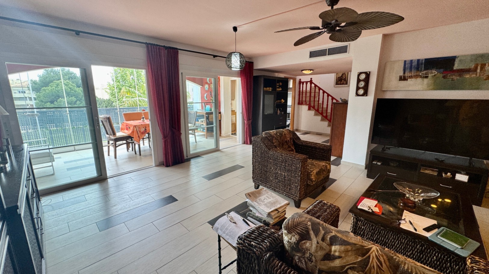 Salon spacieux avec fauteuils en osier, carrelage clair à inserts sombres et grandes baies vitrées ouvrant sur une terrasse. Présence d'un ventilateur de plafond et d'un escalier rouge.