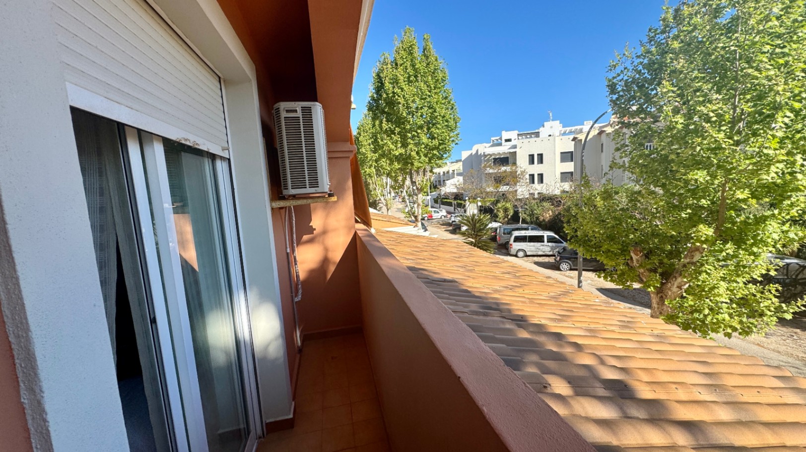 Balcon étroit avec carrelage en terre cuite, unité de climatisation extérieure et vue sur une rue bordée d'arbres et de bâtiments résidentiels.