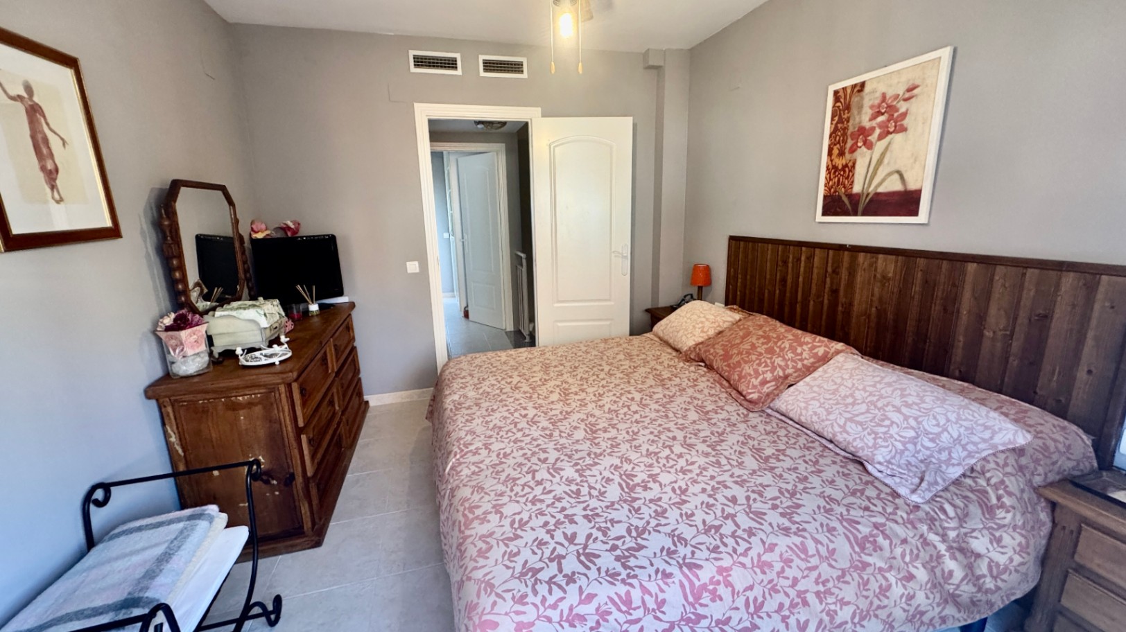 Chambre à coucher avec tête de lit en bois sombre, commode rustique avec miroir et murs gris clair. Sol en carrelage et bouches de climatisation intégrées.