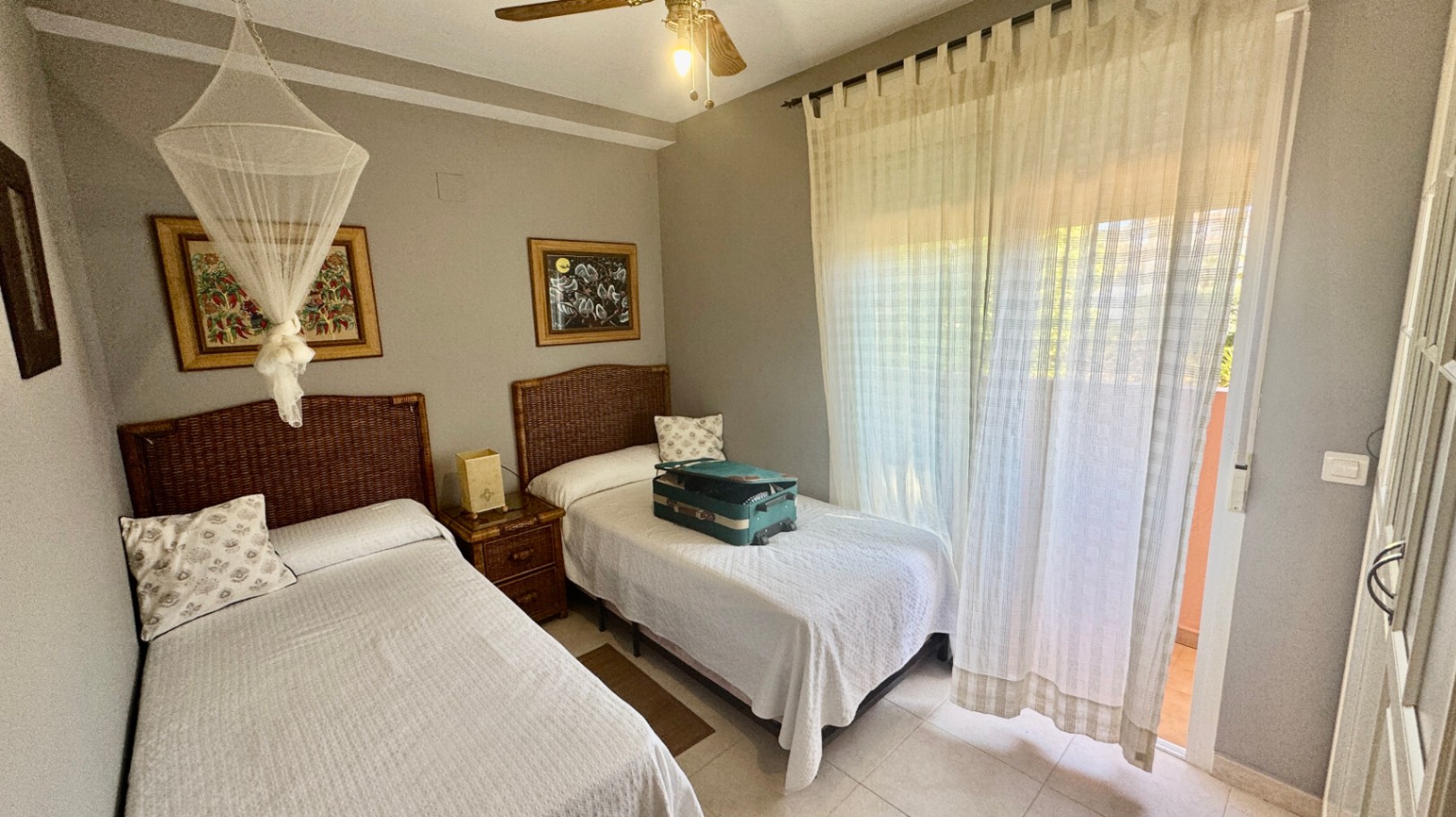 Chambre avec deux lits simples, têtes de lit en osier et accès au balcon par une porte vitrée. La pièce dispose d'un ventilateur de plafond et de carrelage clair.