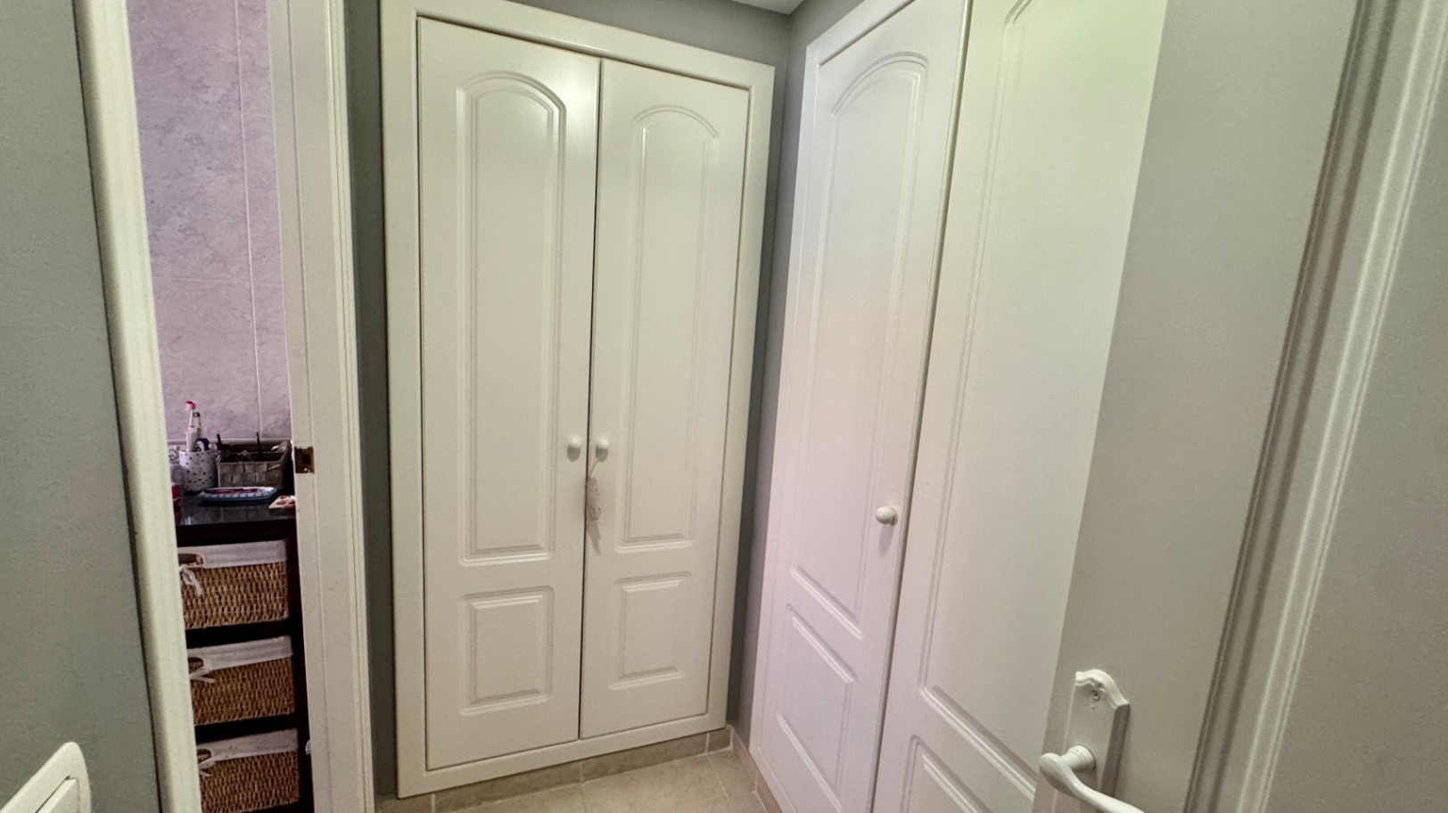 Couloir avec placards intégrés blancs du sol au plafond, dotés de panneaux arqués, situé à côté d'une salle de bain avec rangements en osier.