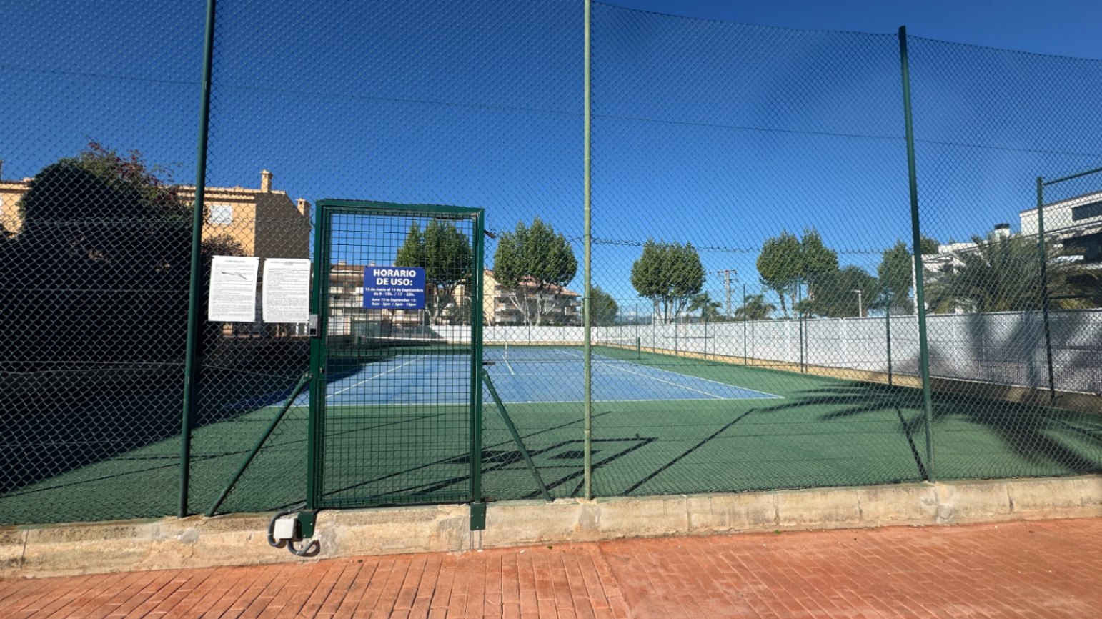 Court de tennis extérieur avec surface dure bleue et verte, entouré d'un grillage de protection élevé et doté d'un portillon d'accès sécurisé.