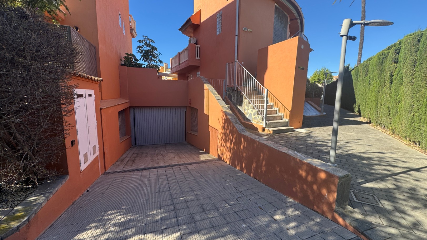 Entrée extérieure avec rampe pavée menant à un garage privé, façade de couleur terre cuite et escalier extérieur avec garde-corps métallique.