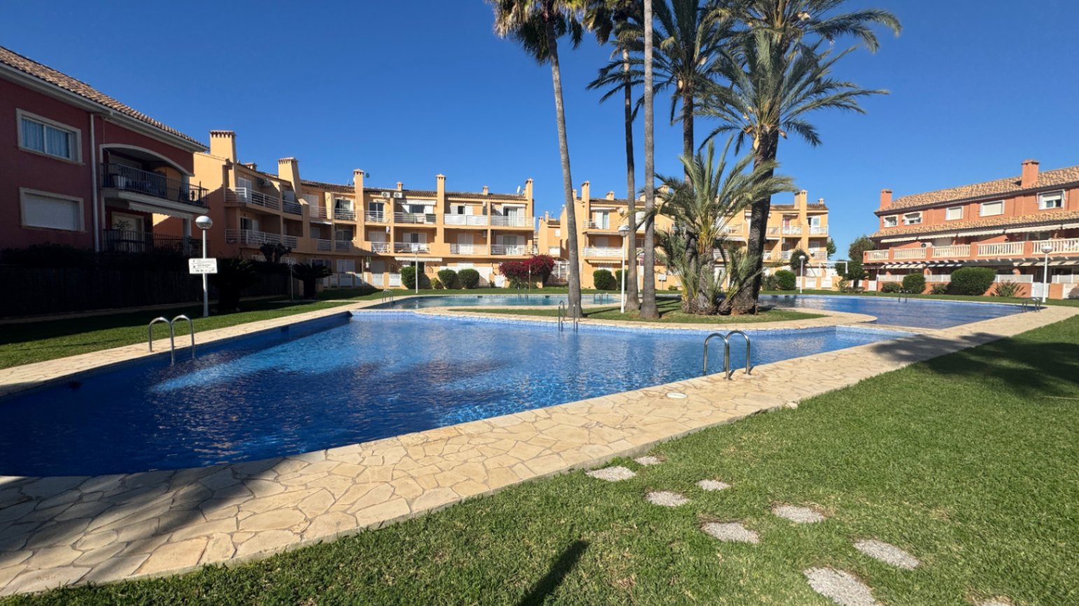Grande piscine communautaire carrelée de bleu, entourée d'une plage en pierre, de pelouses entretenues et de palmiers dans une résidence méditerranéenne.