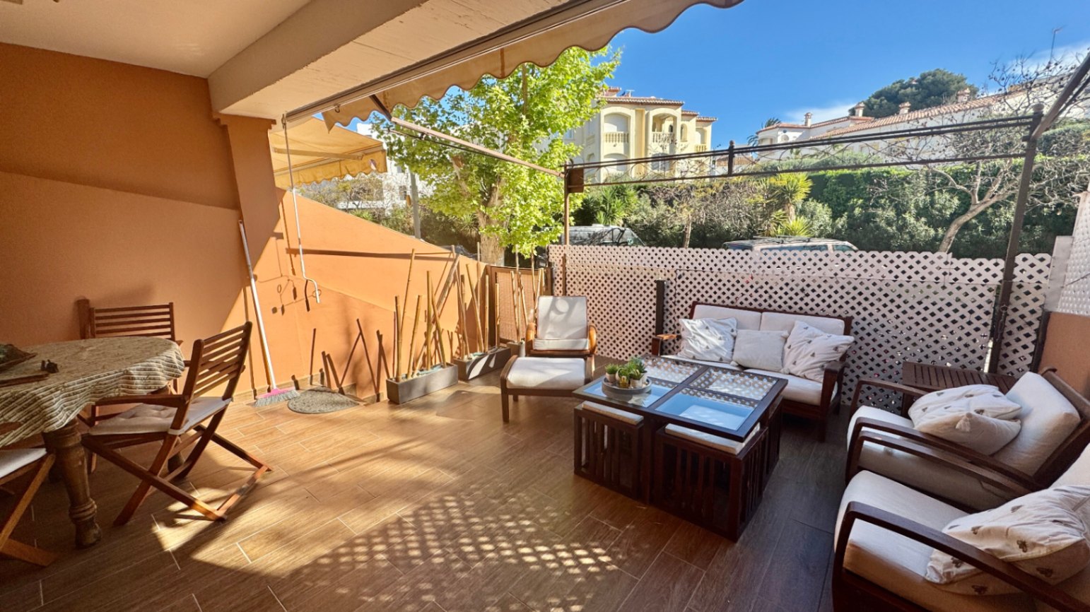 Grande terrasse avec carrelage aspect bois, espace salon avec coussins blancs, coin repas en bois et stores bannes rétractables.