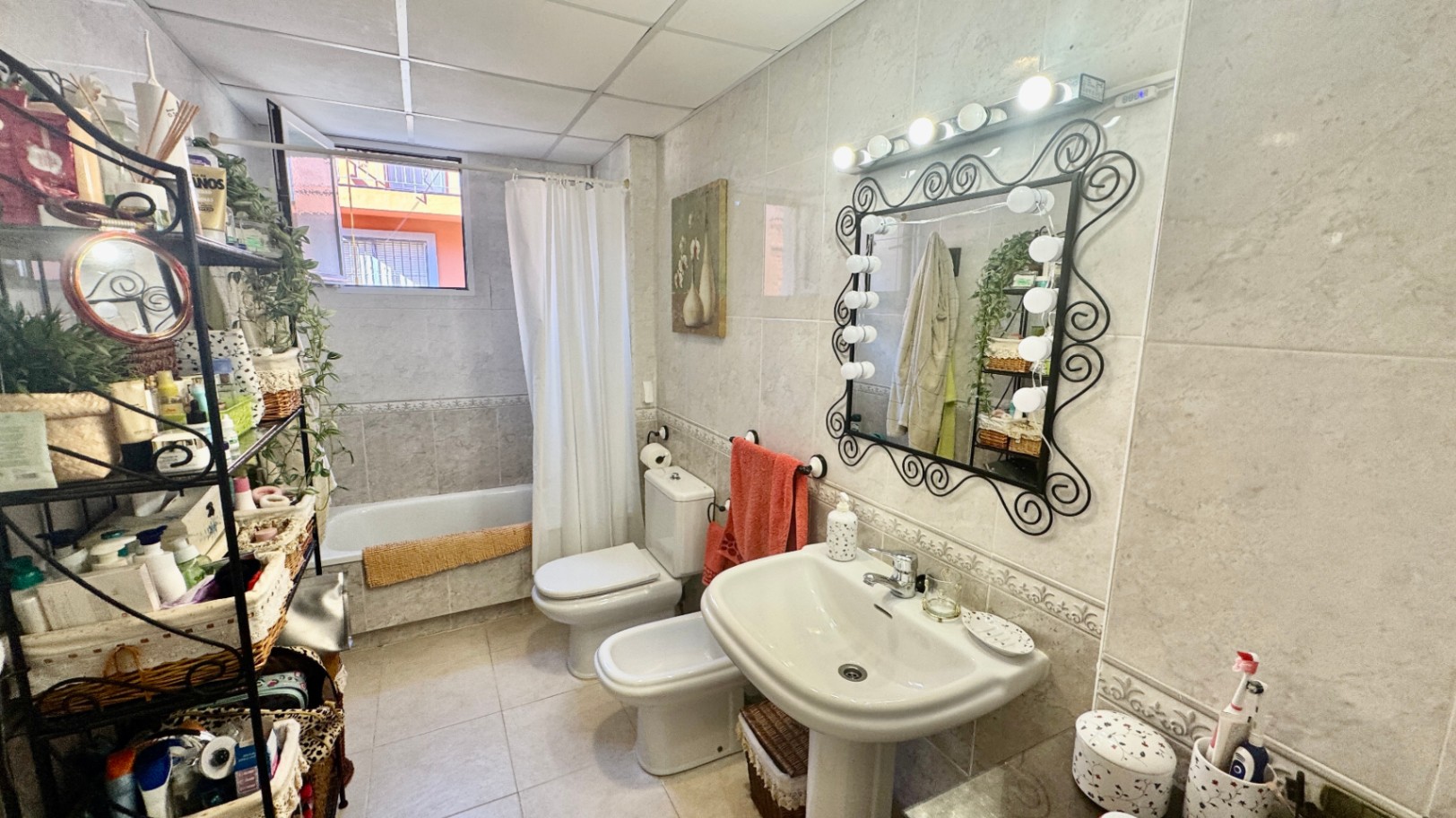 Salle de bain carrelée comprenant un lavabo sur colonne, un miroir en fer forgé avec éclairage style loge, un bidet et une baignoire.