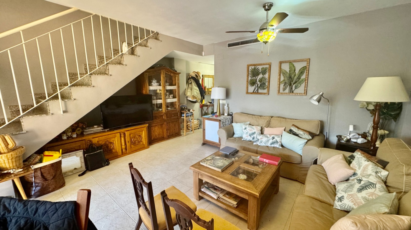 Salon avec canapés beiges, table basse en bois et escalier à rampe blanche. Comprend un meuble TV et un vaisselier en bois intégrés sous l'escalier.