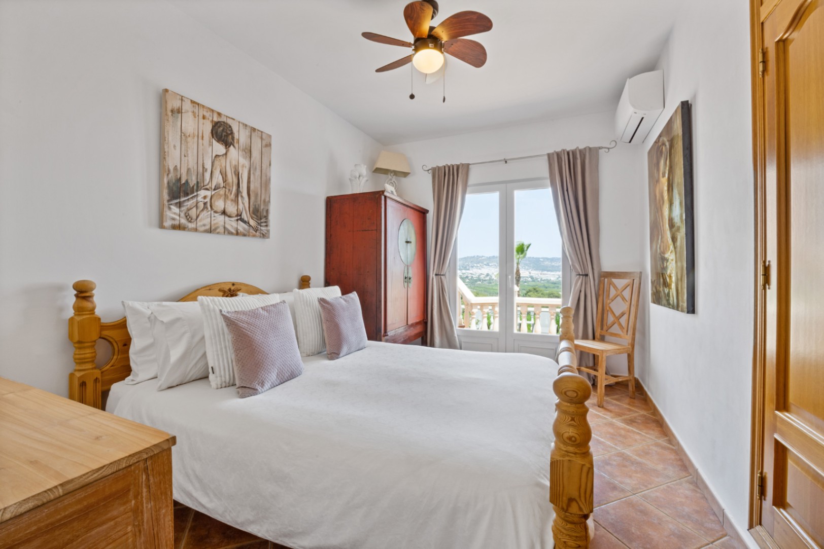 Chambre avec lit en bois, sol en terre cuite et portes-fenêtres ouvrant sur un balcon avec vue. Équipée d'un ventilateur de plafond et de la climatisation.