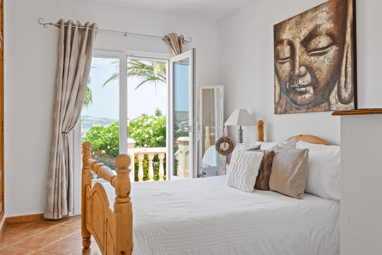 Chambre avec lit en bois, sol en terre cuite et tableau de Bouddha. Une porte-fenêtre s'ouvre sur un balcon offrant une vue sur la végétation méditerranéenne et les collines.