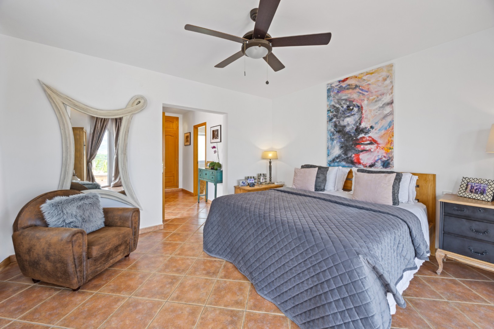 Chambre avec sol en terre cuite, portrait abstrait et miroir décoratif blanc. Elle dispose d'un ventilateur de plafond et d'un fauteuil en cuir avec coussin en fourrure.