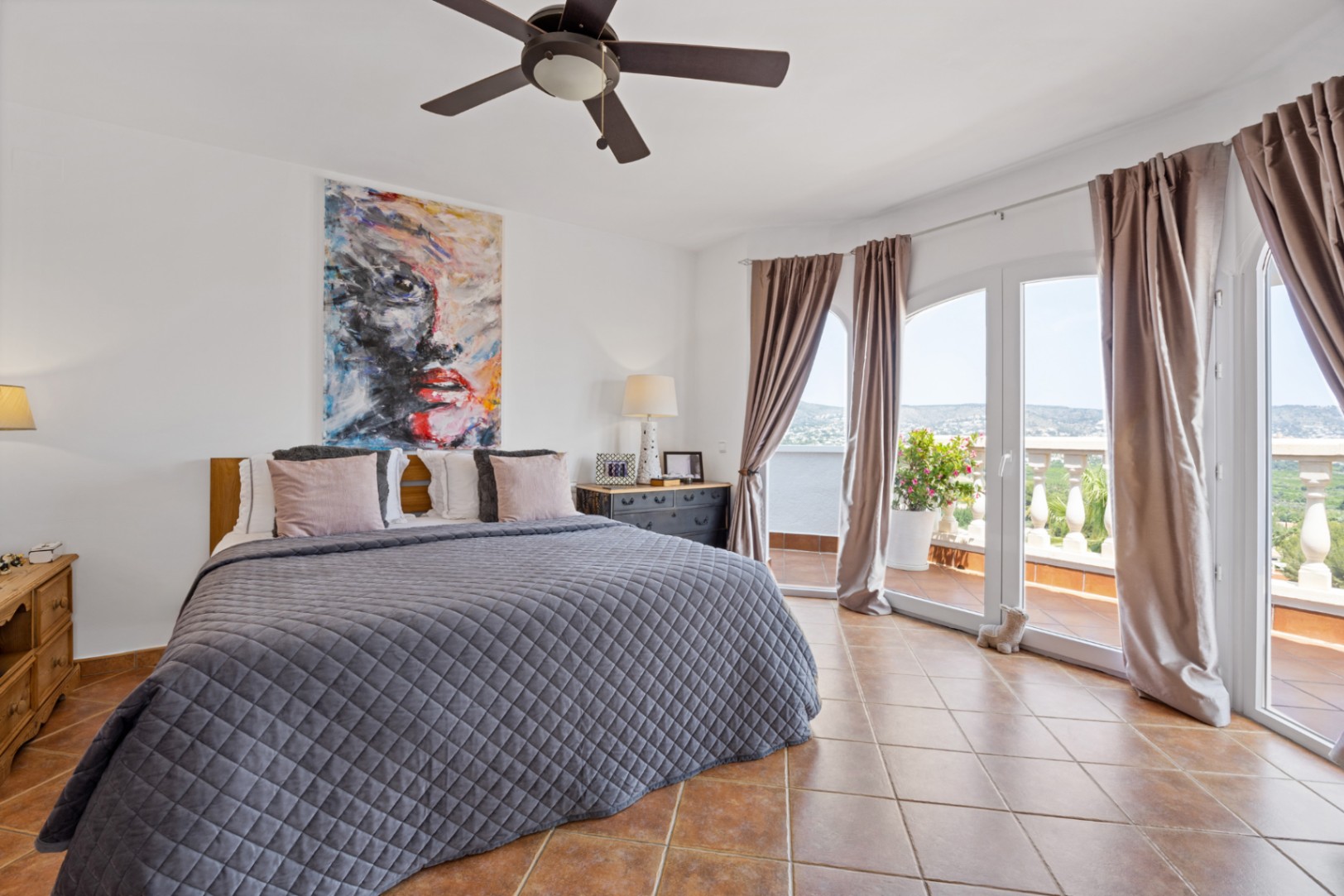 Chambre avec sol en terre cuite, ventilateur de plafond et portes vitrées cintrées s'ouvrant sur une terrasse avec vue sur la vallée.