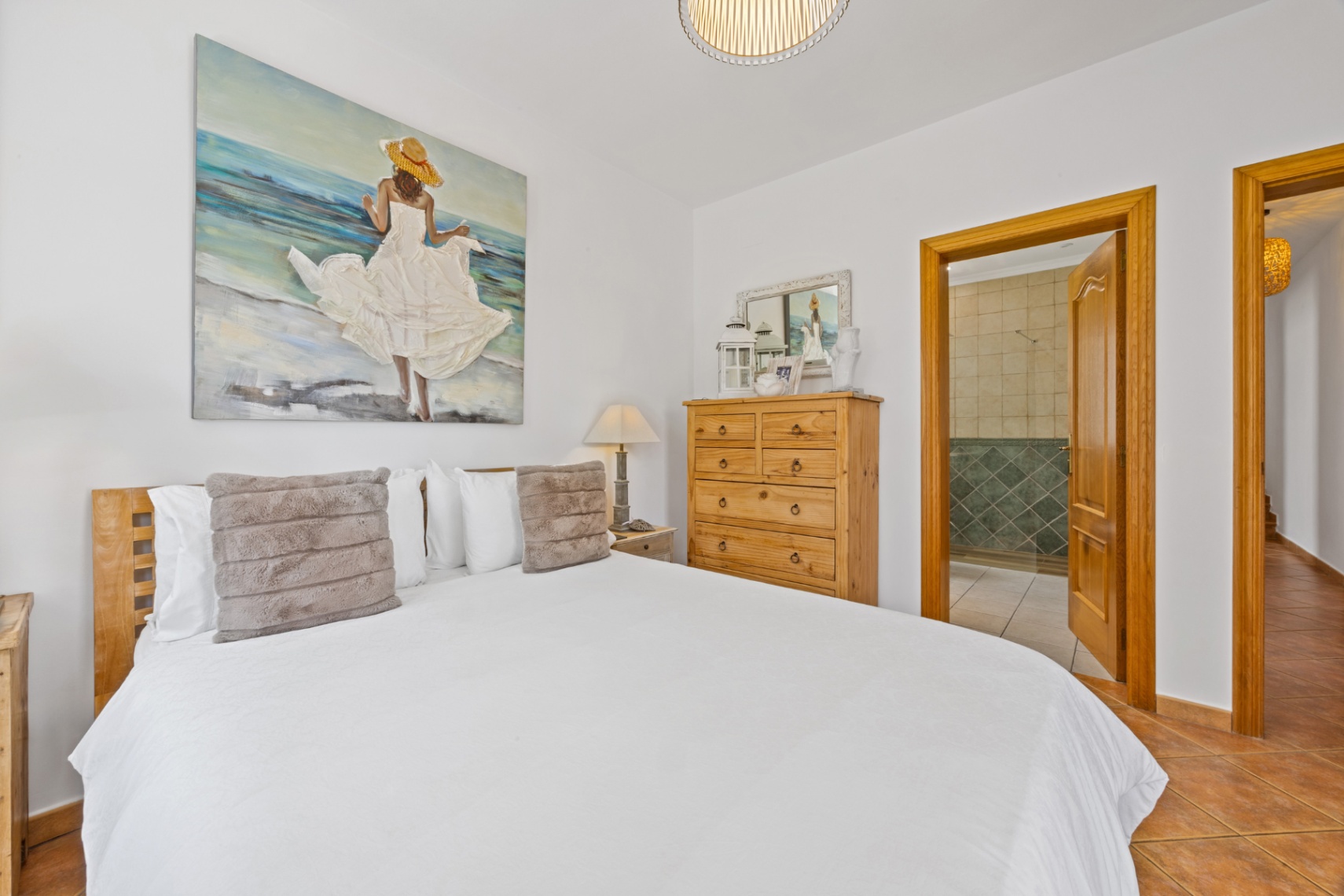 Chambre avec tête de lit en bois, peinture murale côtière et sol en terre cuite. Une porte ouverte mène à une salle de bain attenante carrelée en vert.