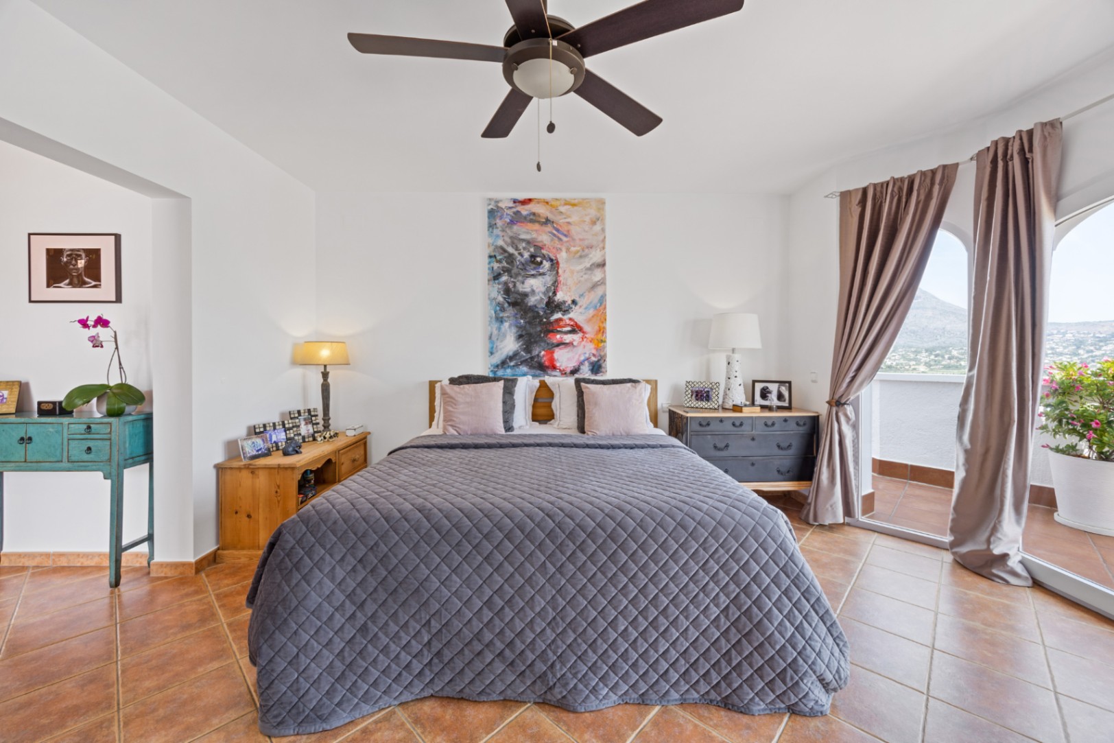 Chambre lumineuse dotée d'un sol en terre cuite, d'un ventilateur de plafond et de baies vitrées ouvrant sur un balcon privé avec vue sur la montagne.