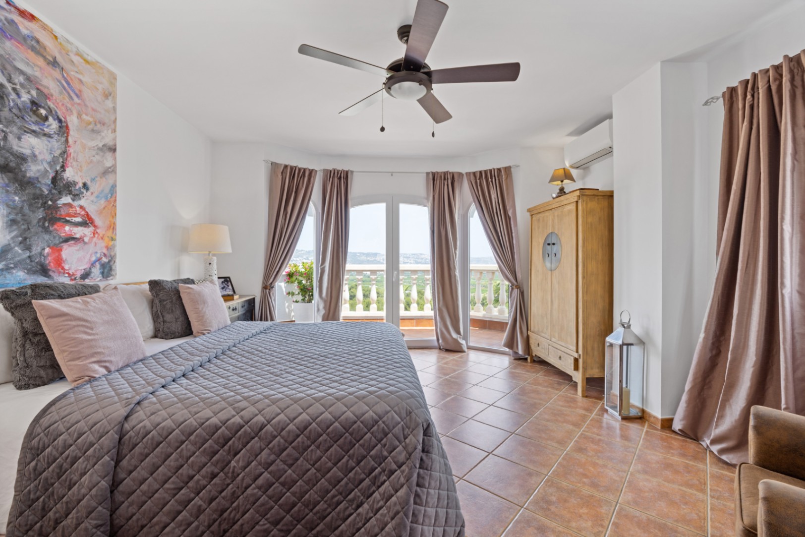 Chambre spacieuse avec sol en terre cuite, ventilateur de plafond et portes-fenêtres cintrées ouvrant sur un balcon offrant une vue panoramique.