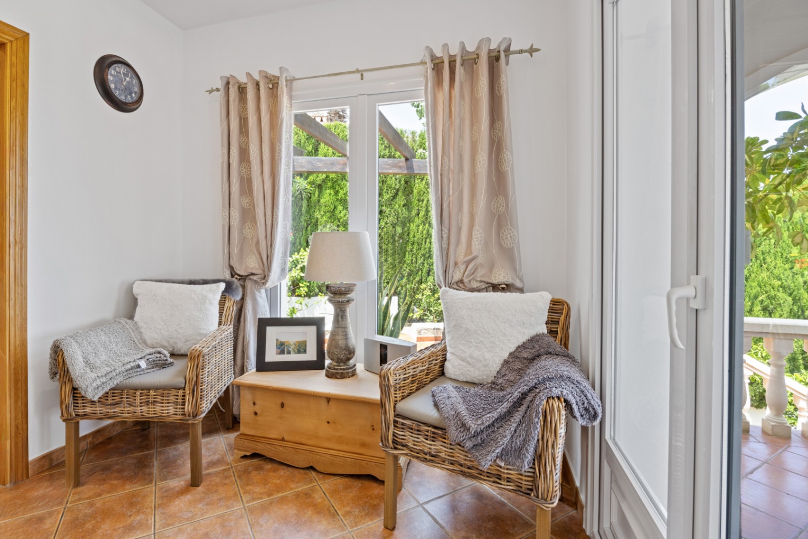 Coin salon avec deux fauteuils en osier, coffre en bois rustique et sol en terre cuite. Les fenêtres offrent une vue sur le jardin et la pergola extérieure.