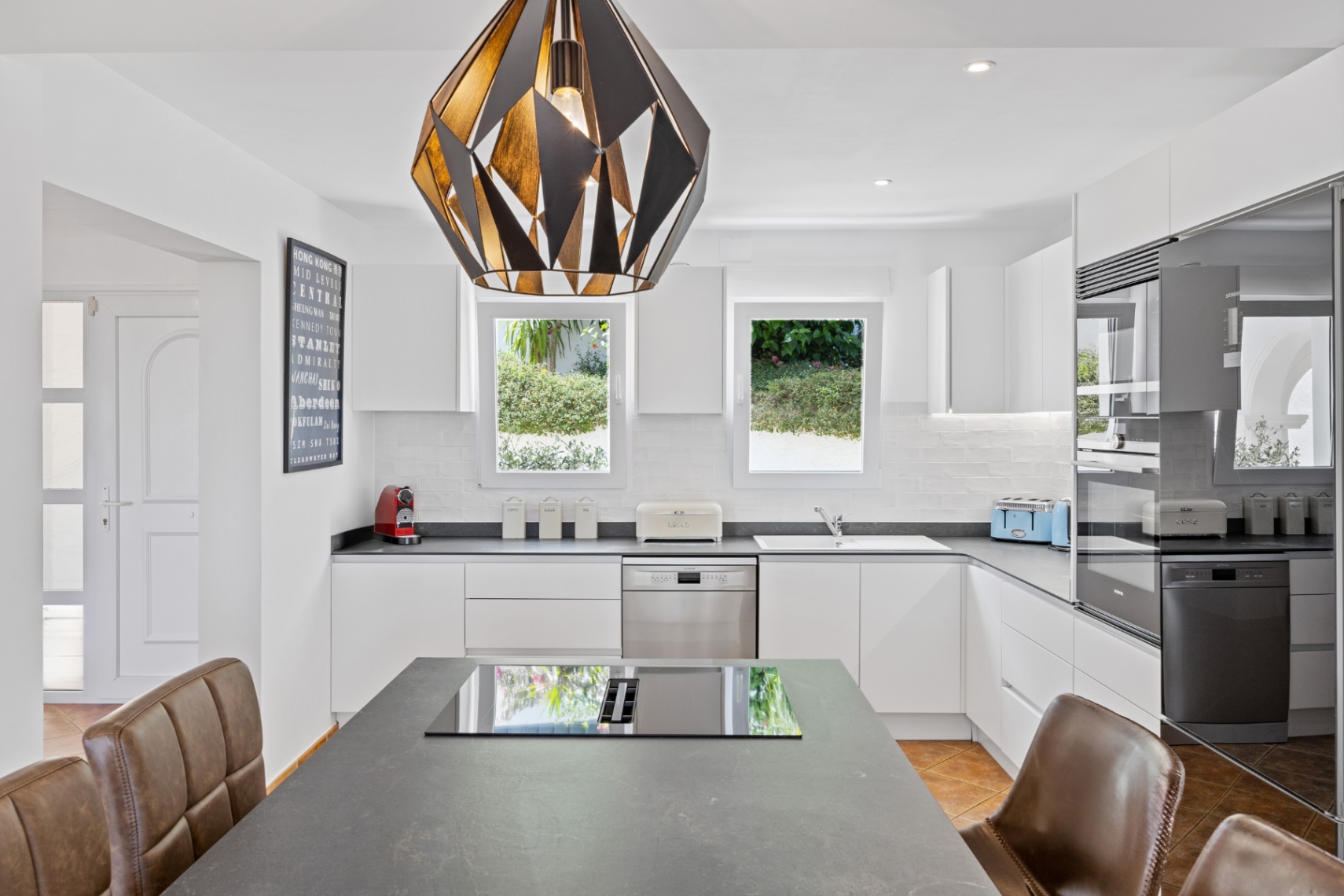 Cuisine moderne aux finitions blanches mates, plans de travail en pierre grise et îlot central avec plaque de cuisson, éclairé par une suspension géométrique.