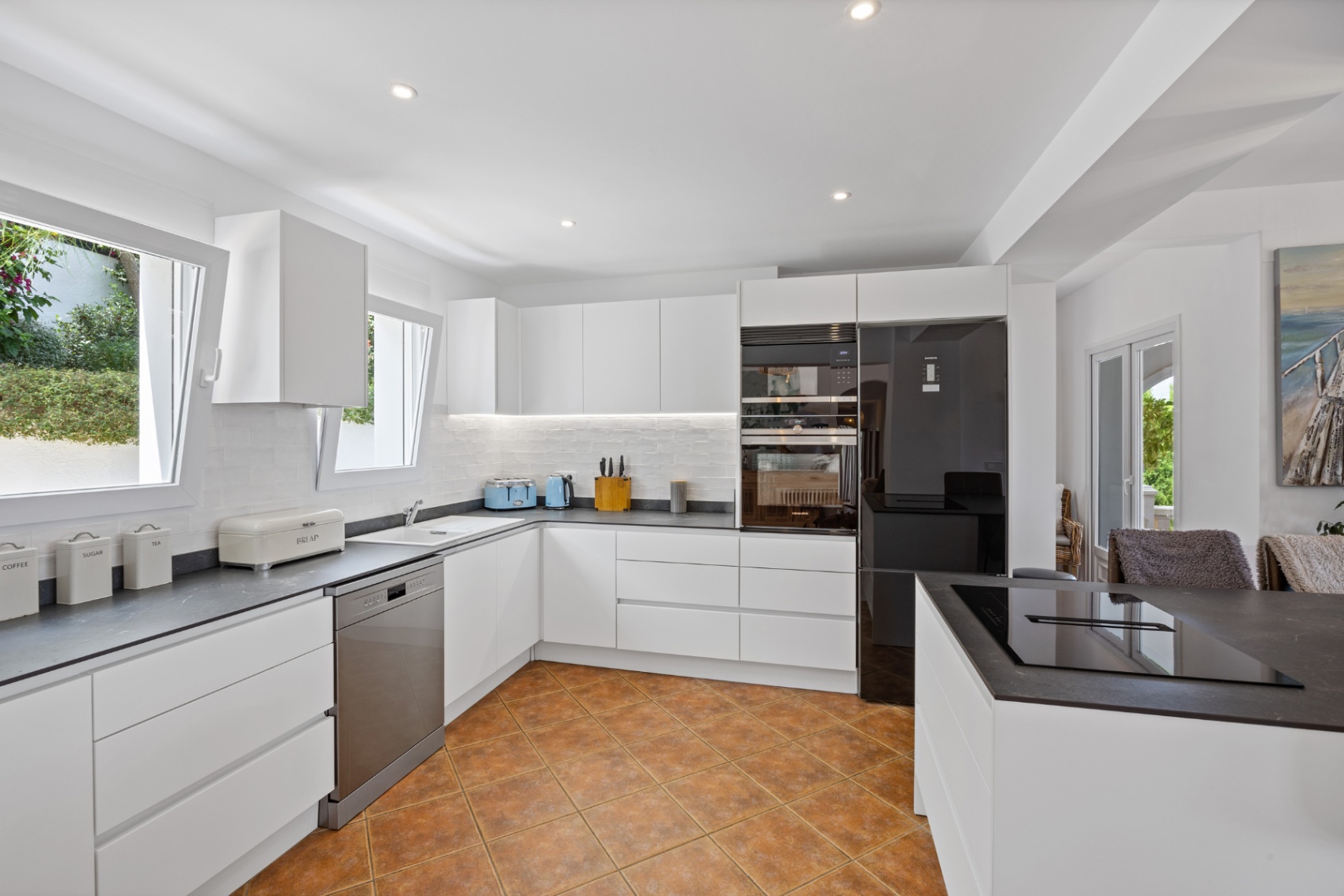 Cuisine moderne avec placards blancs sans poignées, plans de travail sombres effet pierre, appareils intégrés et sol en terre cuite céramique.