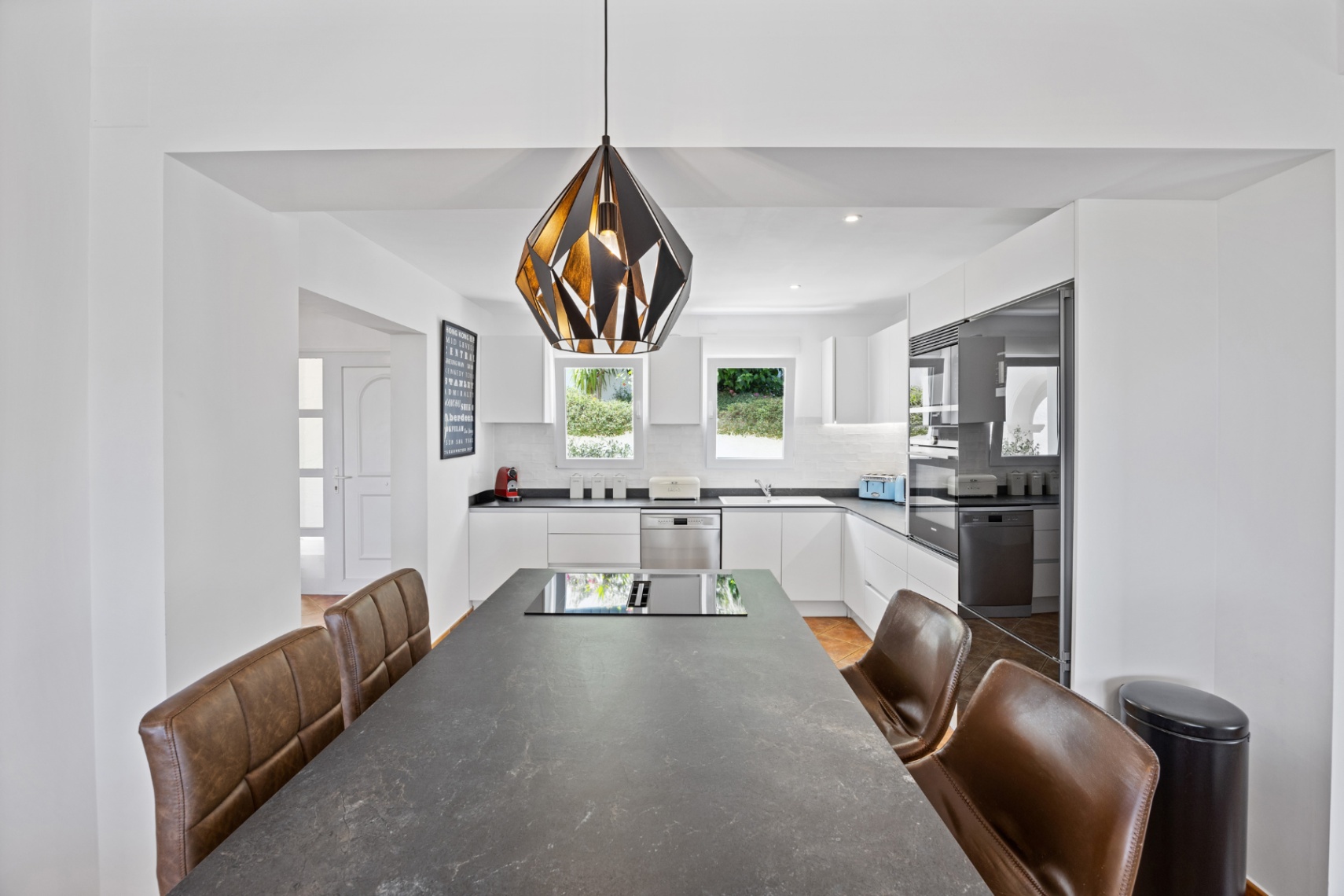 Cuisine moderne avec un grand îlot en pierre sombre, plaque à induction intégrée, chaises en cuir marron et meubles blancs avec électroménager encastré.