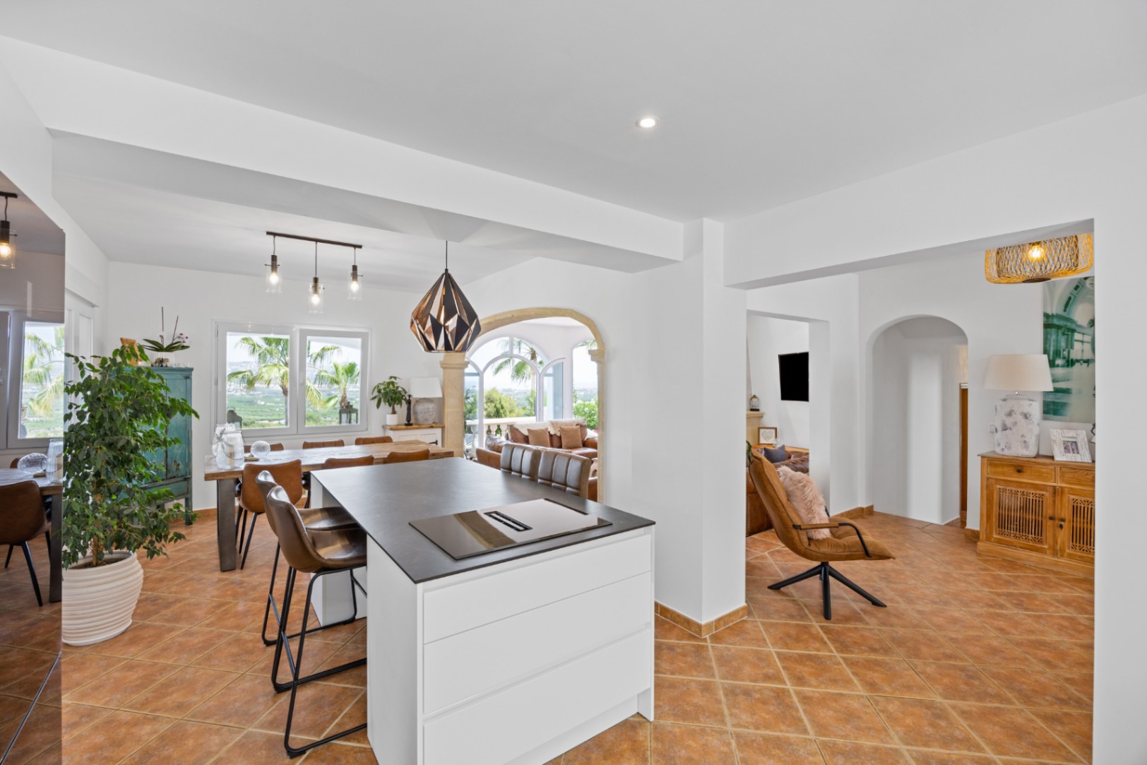 Cuisine ouverte avec îlot blanc et plan de travail sombre, sol en terre cuite et coin repas offrant une vue sur les palmiers à travers de grandes fenêtres.