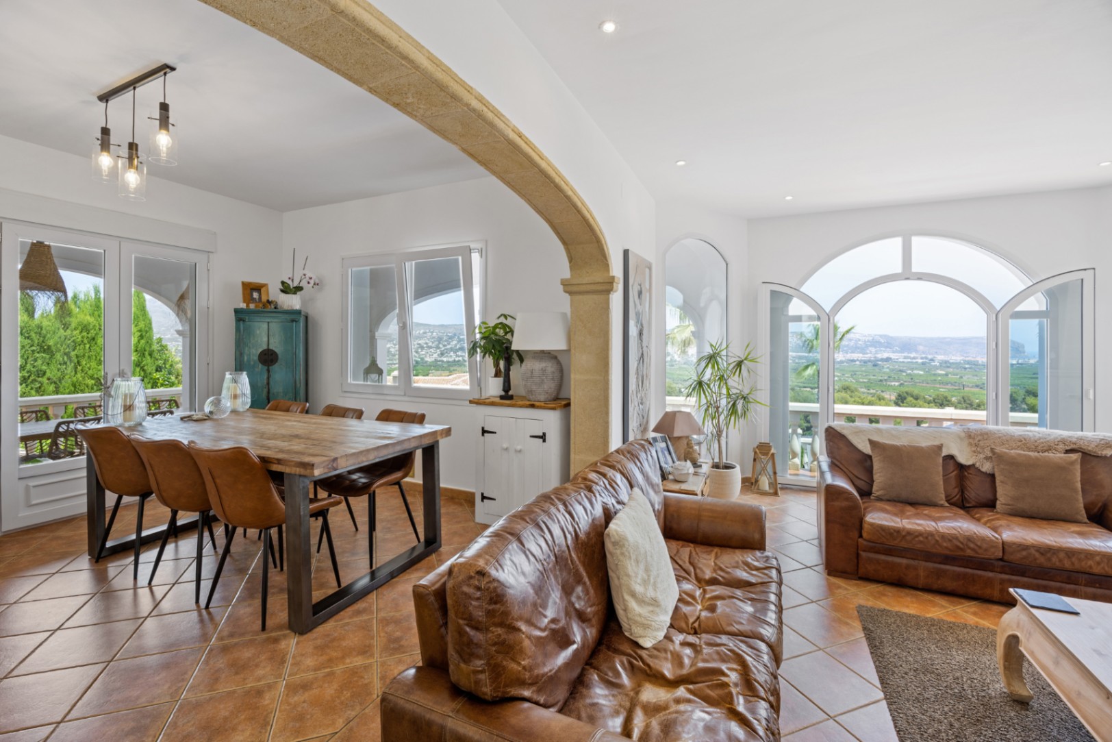 Espace de vie ouvert avec arche en pierre, canapés en cuir et table en bois. Les portes cintrées offrent une vue panoramique sur la vallée environnante.