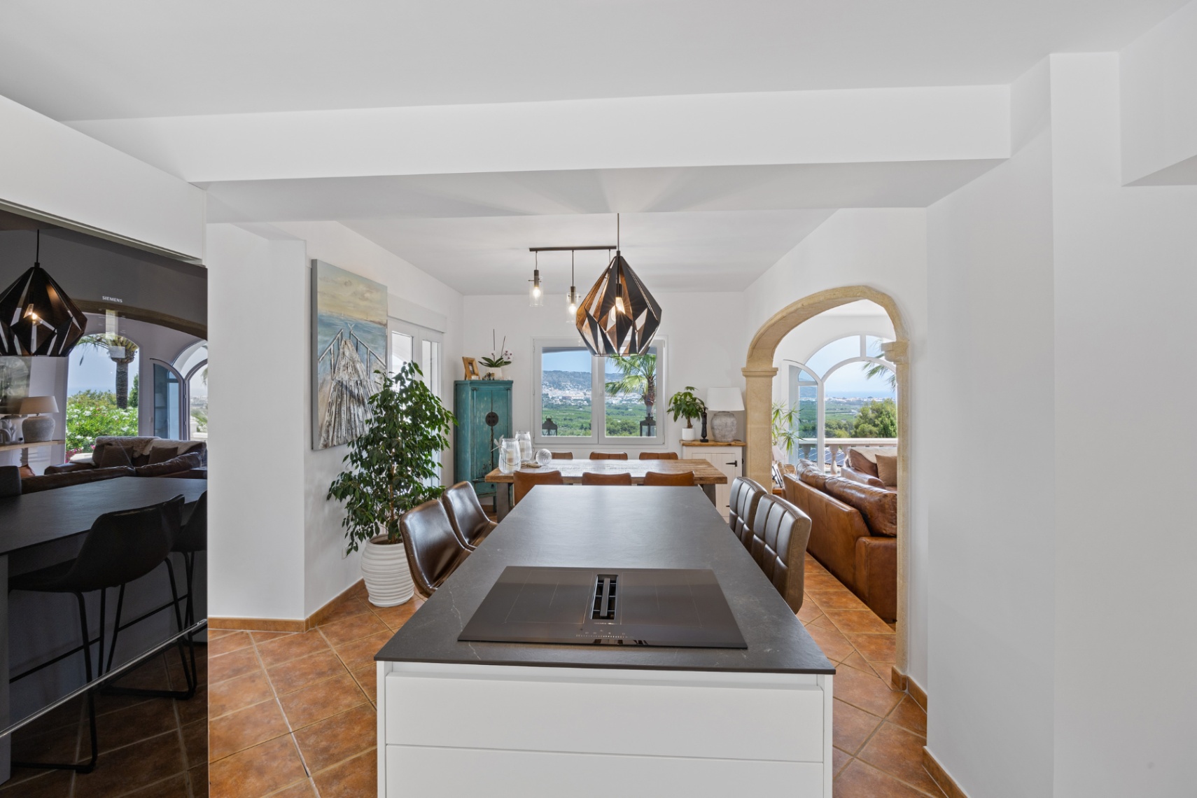 Îlot de cuisine moderne avec plaque à induction et hotte intégrée, sol en terre cuite et arche en pierre ouvrant sur un salon avec vue panoramique sur la vallée.