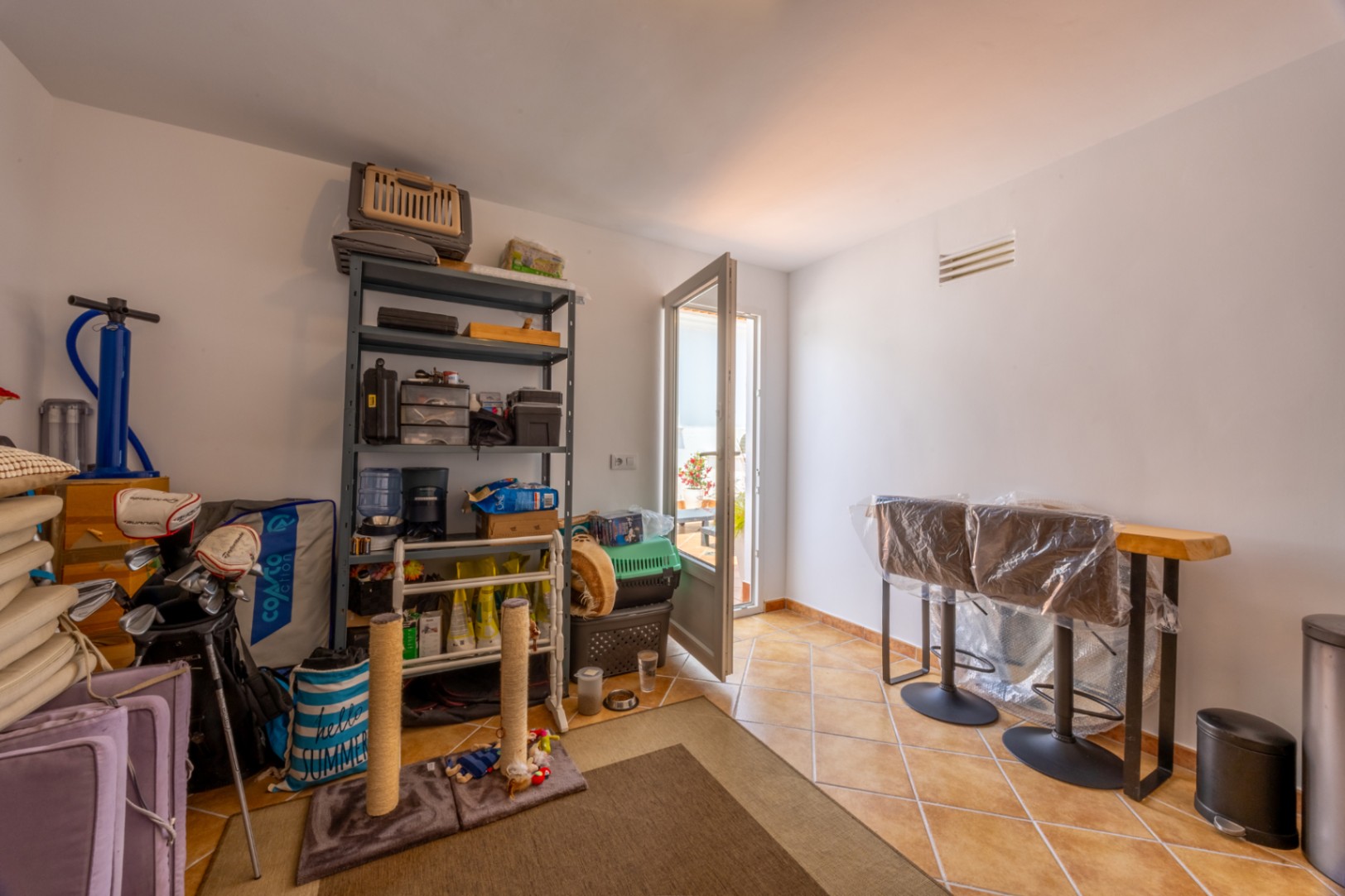 Pièce de rangement avec sol en terre cuite, comprenant une étagère métallique, une table haute en bois avec tabourets et un accès vers la terrasse.