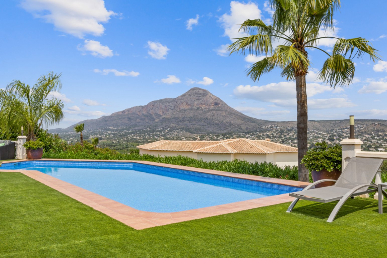 Piscine extérieure avec mosaïque bleue, entourée d'un gazon synthétique et d'une bordure en terre cuite, offrant une vue panoramique sur un sommet montagneux.