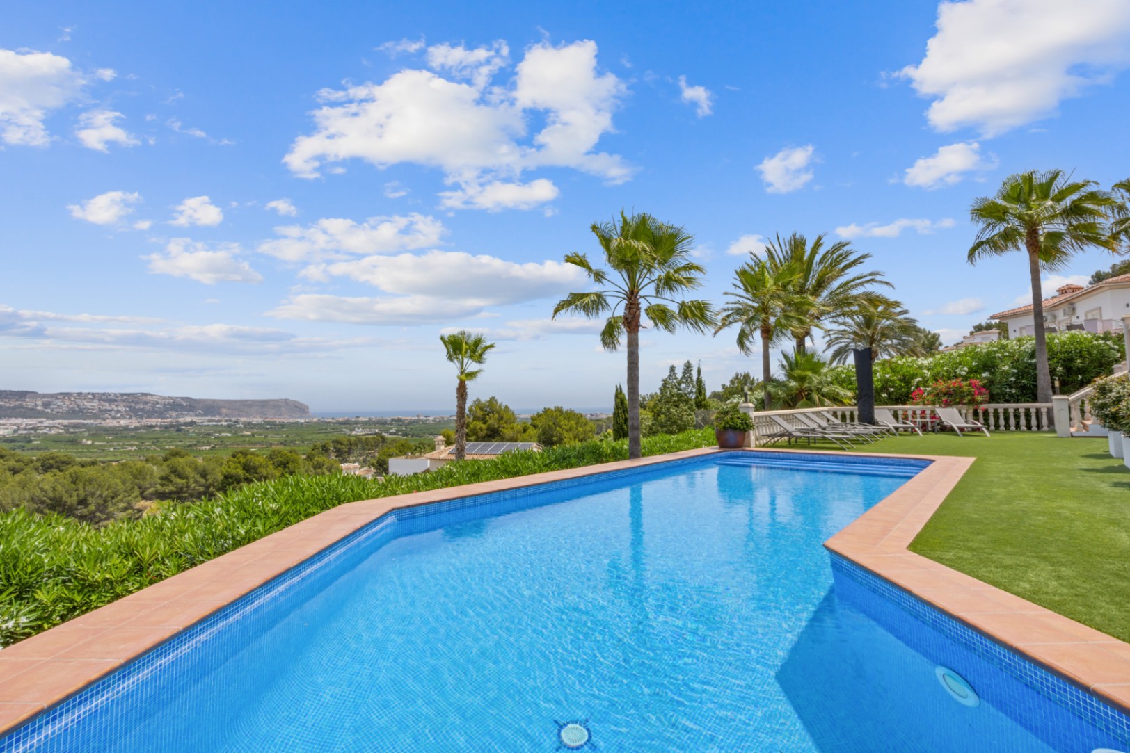 Piscine extérieure avec mosaïque bleue et bordure en terre cuite, offrant une vue panoramique sur la vallée et la mer, entourée de palmiers et d'un gazon.