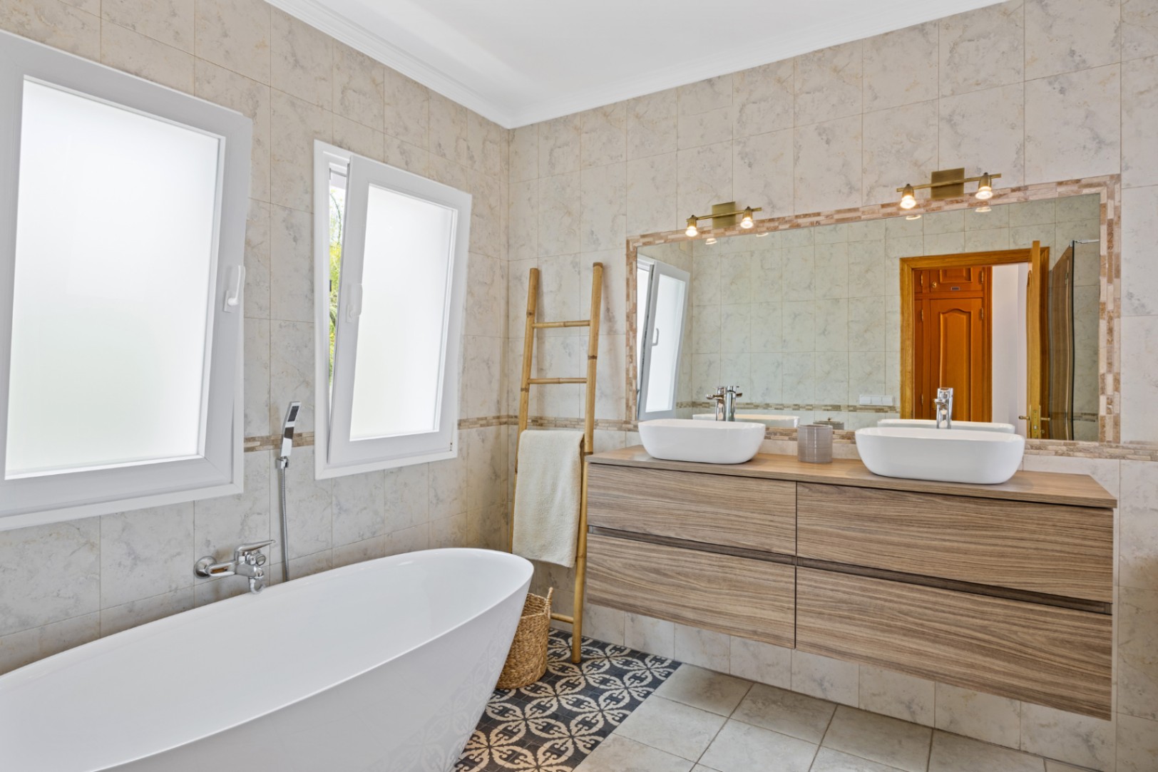 Salle de bain avec baignoire îlot, double vasque sur meuble en bois et sol en carreaux de ciment. Grand miroir avec luminaires en laiton et fenêtres à vitrage dépoli.