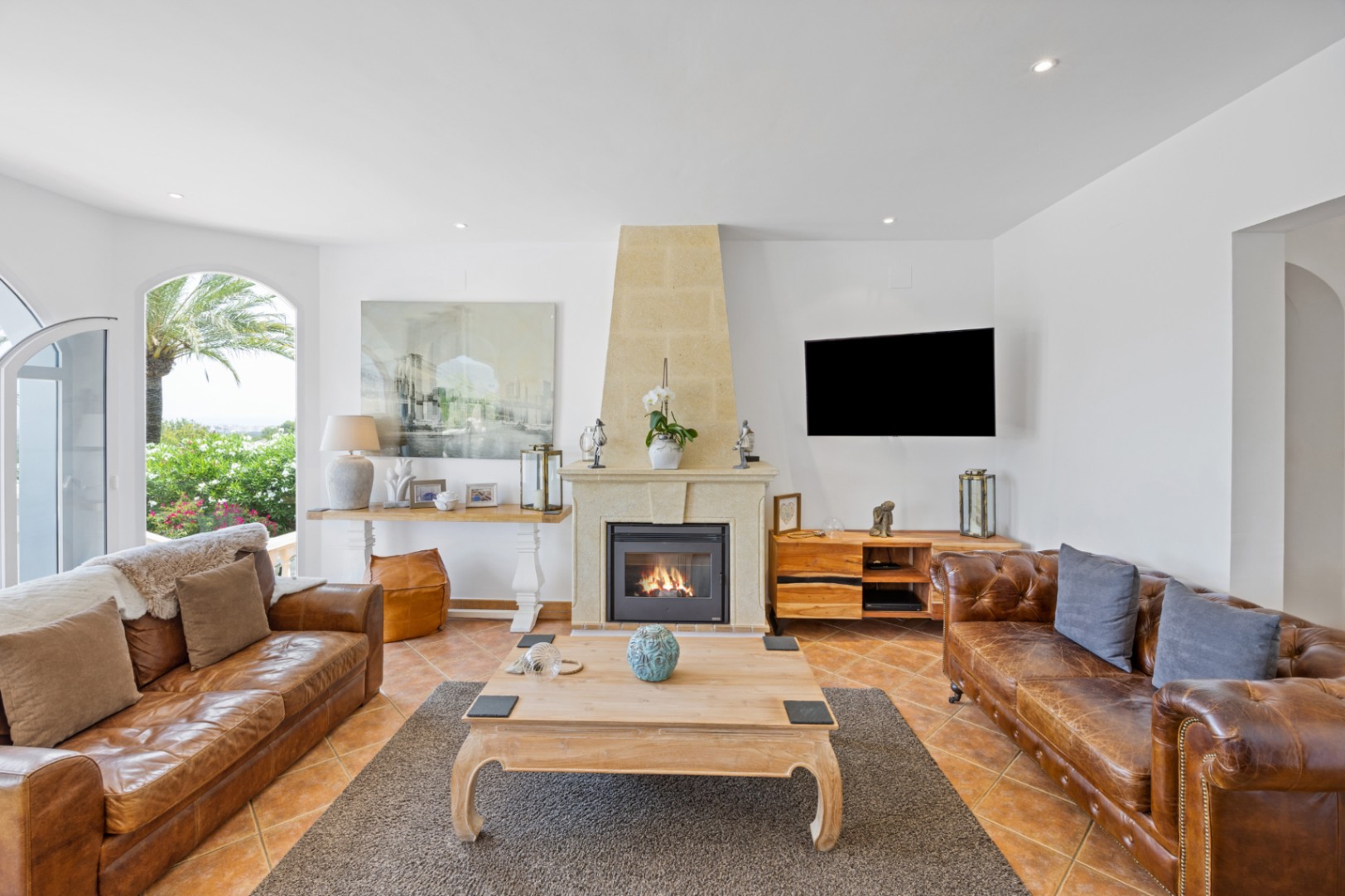 Salon élégant avec cheminée en pierre, canapés en cuir marron et sol en terre cuite. Une porte cintrée vitrée s'ouvre sur un jardin arboré de palmiers.