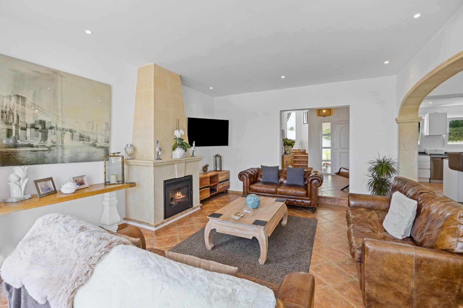 Salon spacieux avec canapés en cuir marron, cheminée en pierre et sol en terre cuite. Une arche en pierre sculptée s'ouvre sur la cuisine et l'entrée.