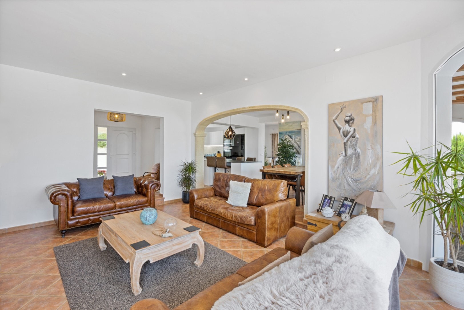 Salon spacieux avec canapés en cuir marron, sol en terre cuite et arche en pierre menant à la salle à manger et à la cuisine ouverte.